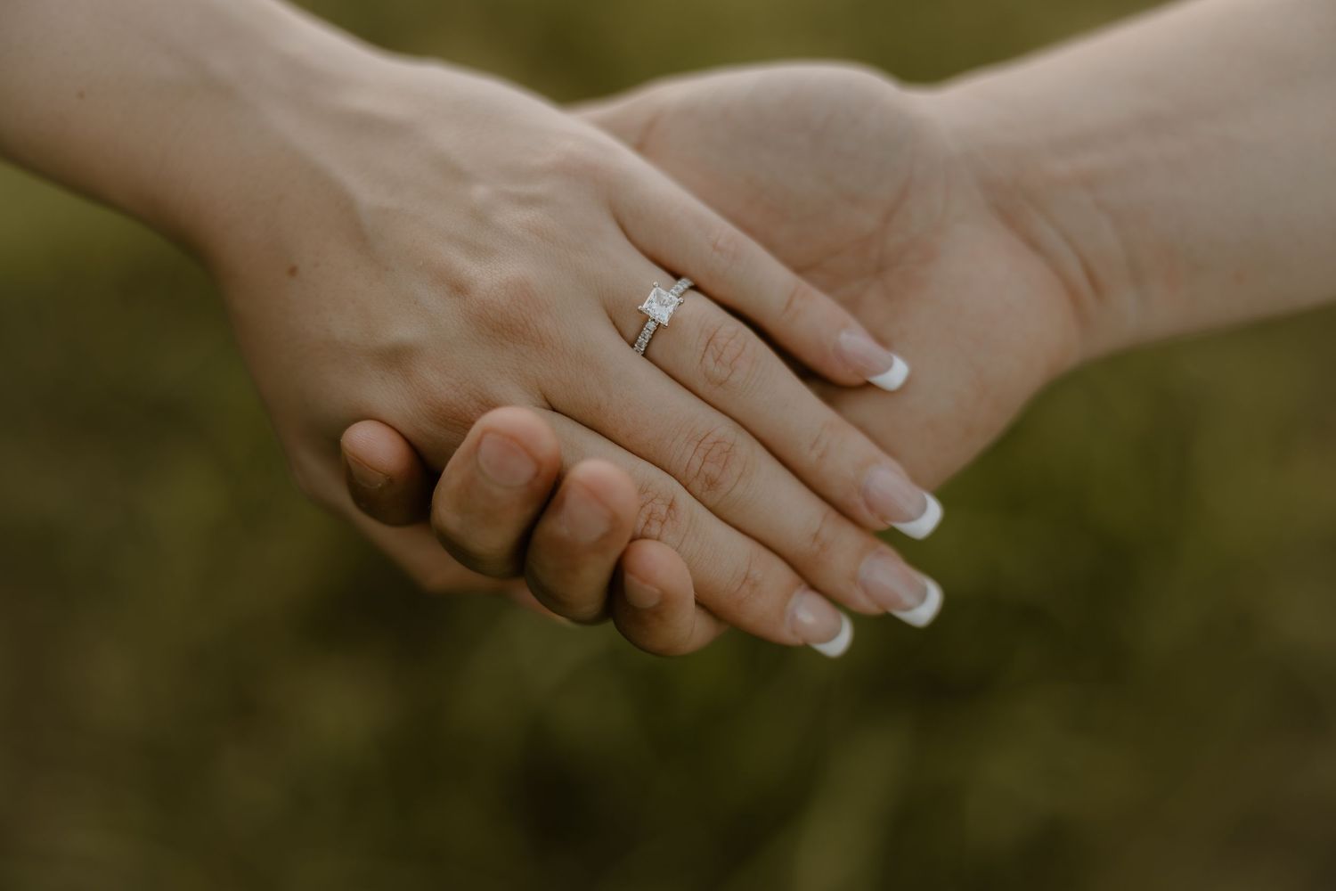 Two hands clasped together against a blurred green background, with an engagement ring visible on one hand.
