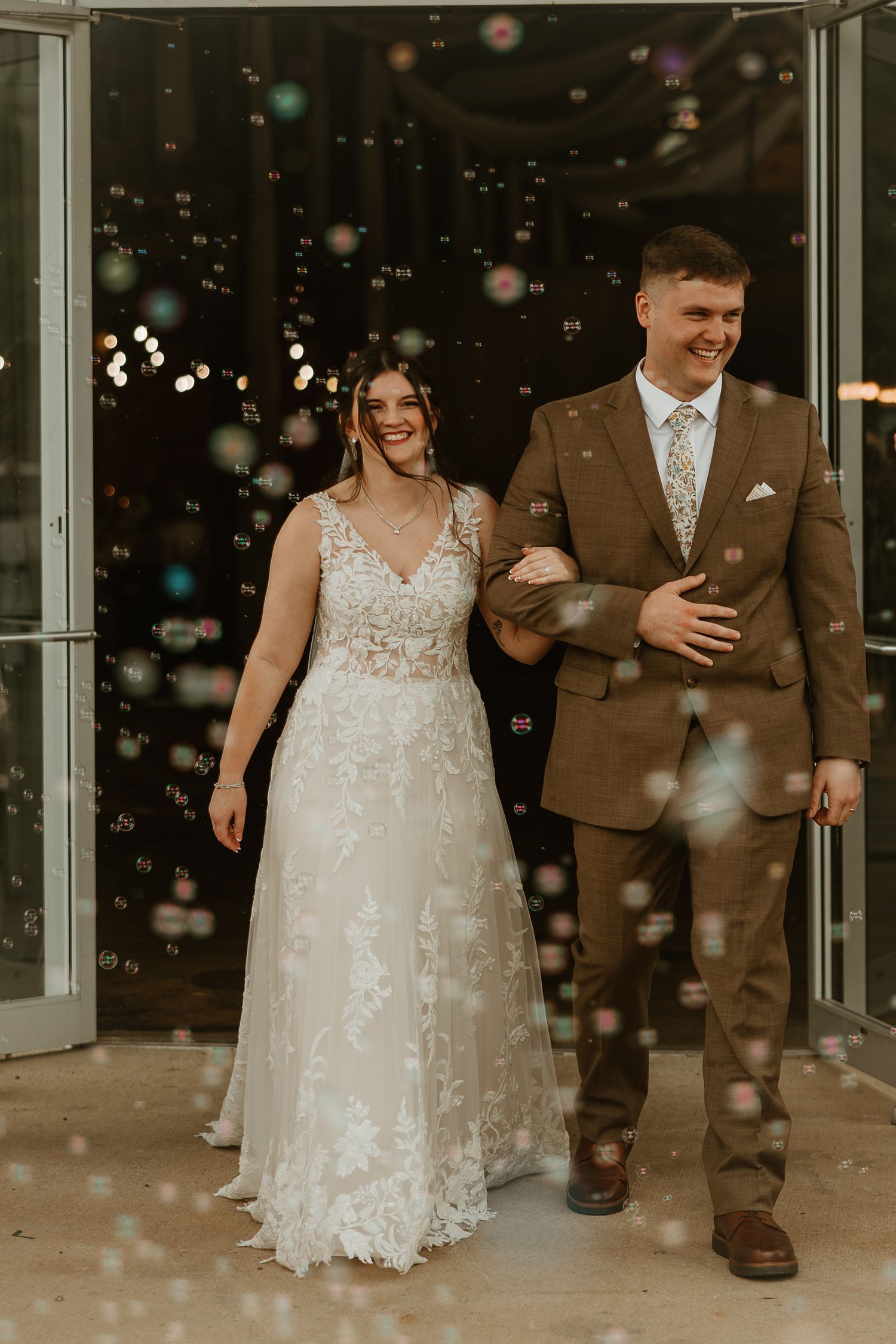 Newlyweds exit venue together amid floating bubbles while wearing brown suit and lace wedding gown.