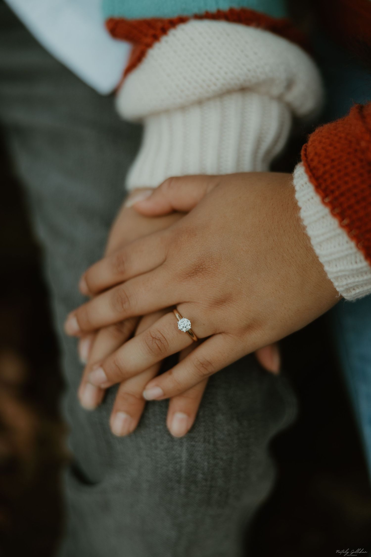 Close-up view of hands wearing red and white knit sweater sleeves with delicate engagement ring on finger.