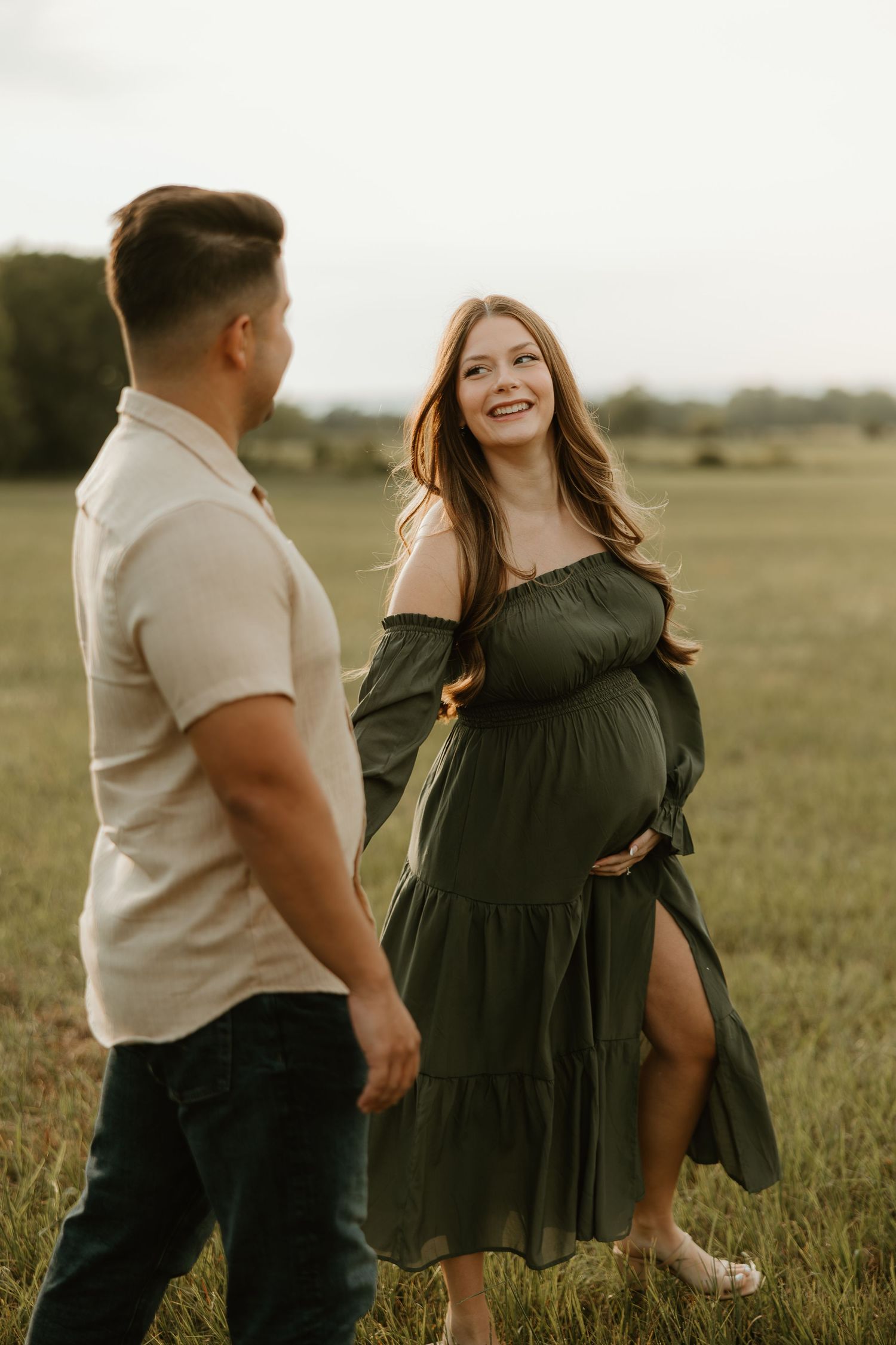 A couple in casual attire dance together playfully in a sunlit grassy field during golden hour.