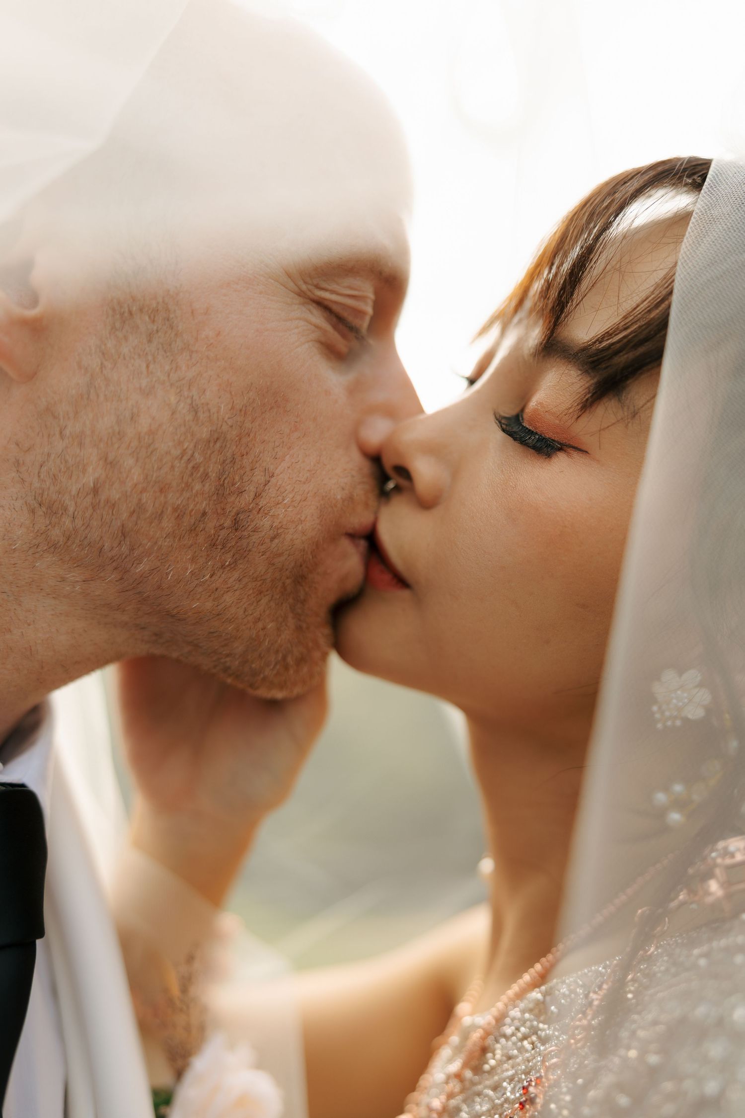 A bride and groom share a romantic kiss under a flowing white wedding veil on their wedding day.