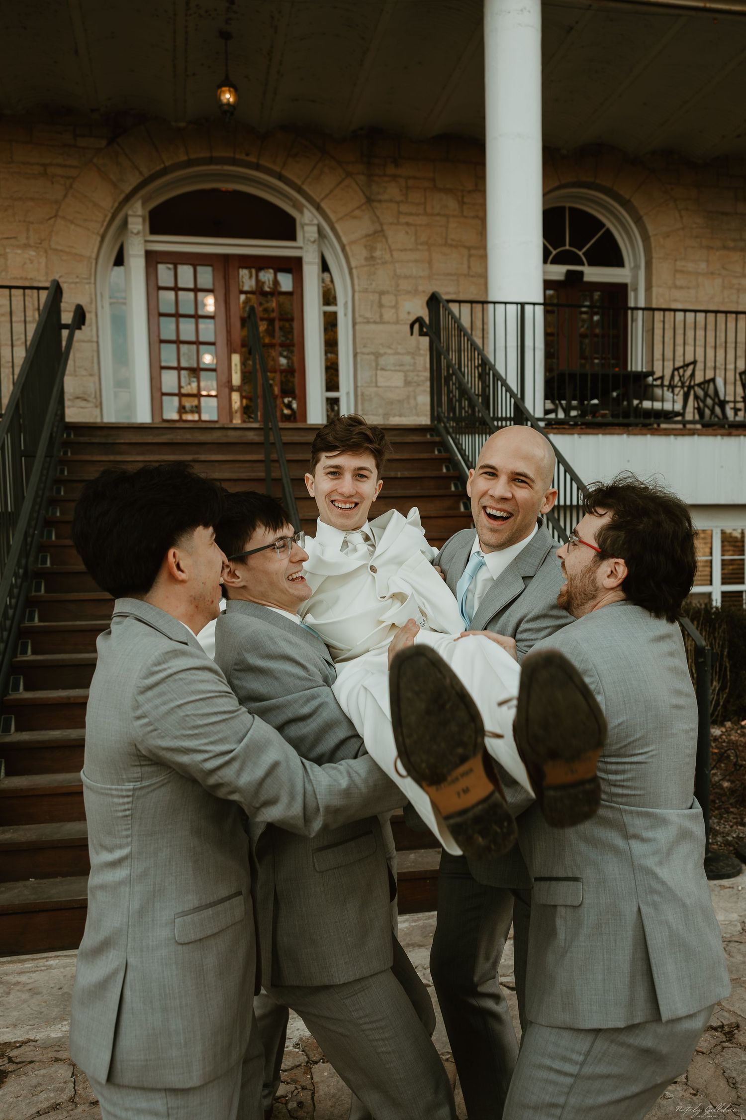 Groomsmen in matching grey suits playfully lift someone in white attire on stone steps of a historic building.