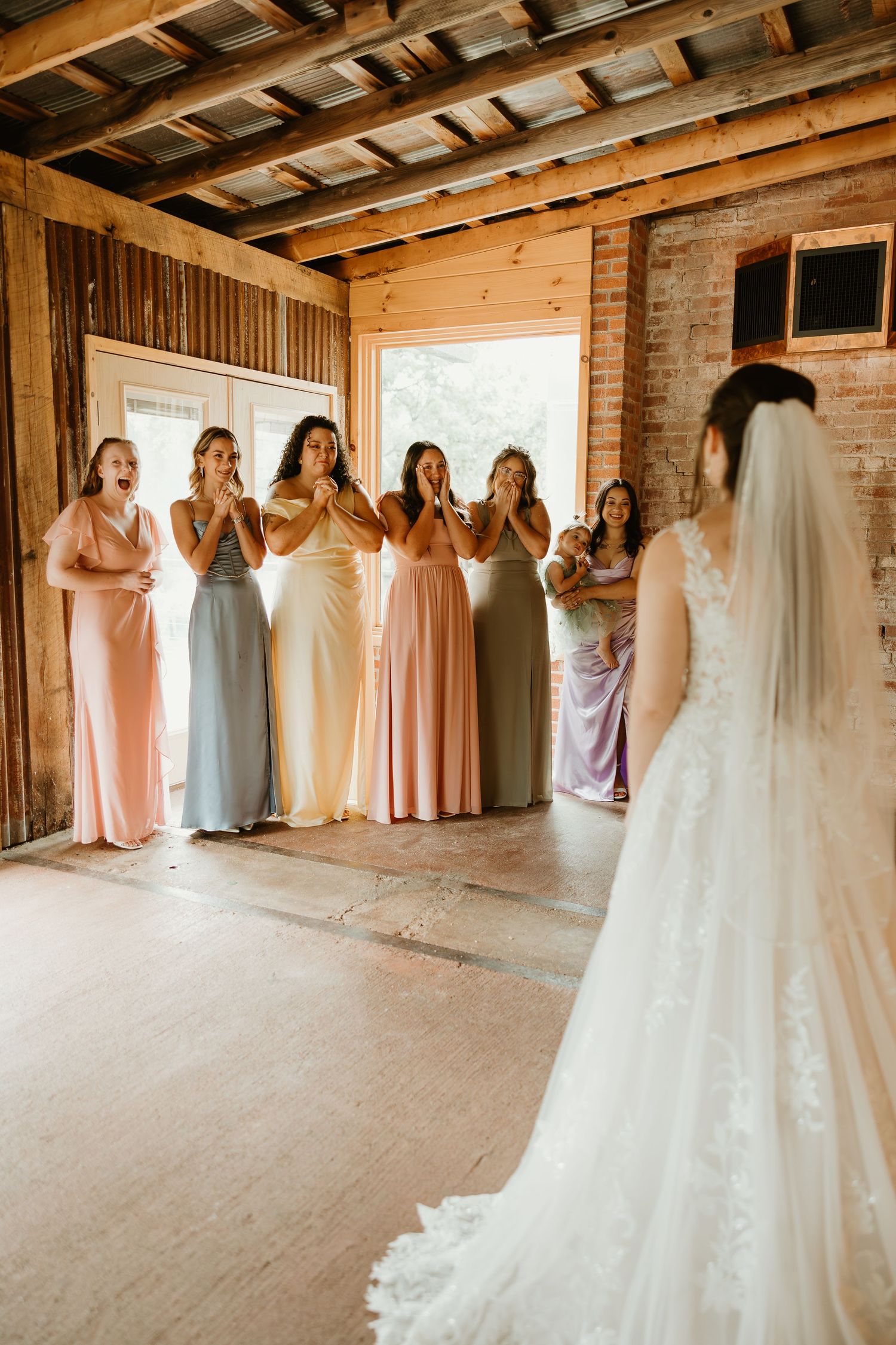 Bridesmaids in soft pastel dresses react emotionally to seeing bride in white lace gown at rustic barn venue.