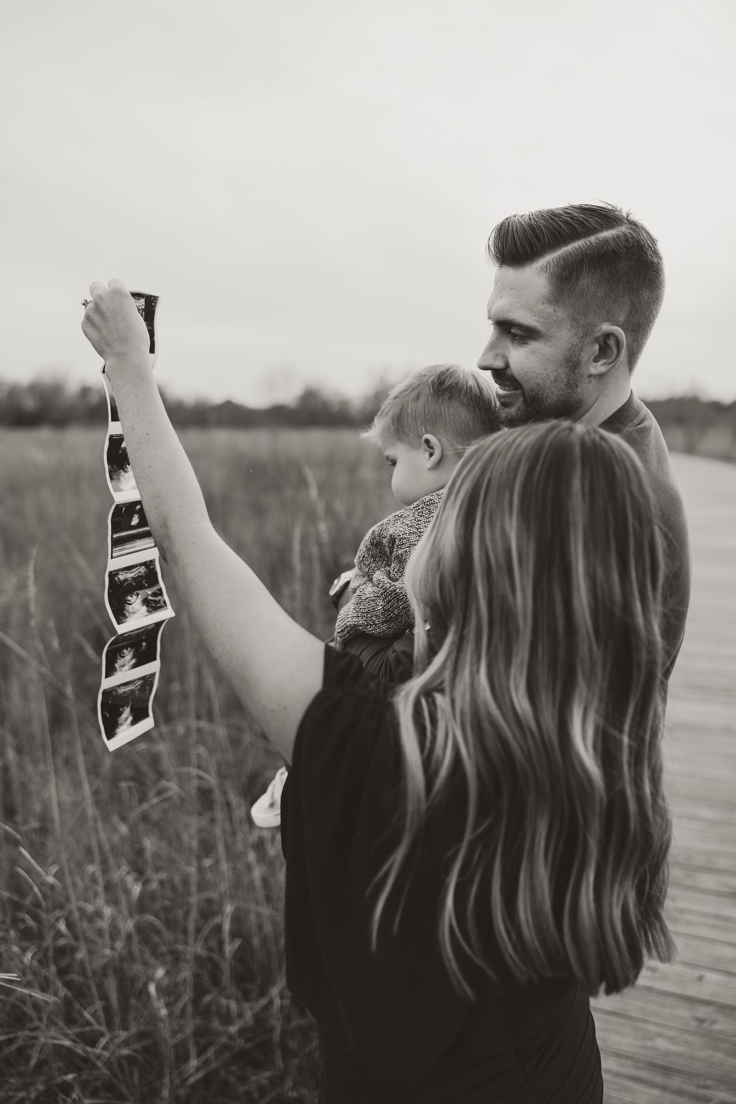 A couple stands together in a field holding up a sonogram photo in a series of black and white images.