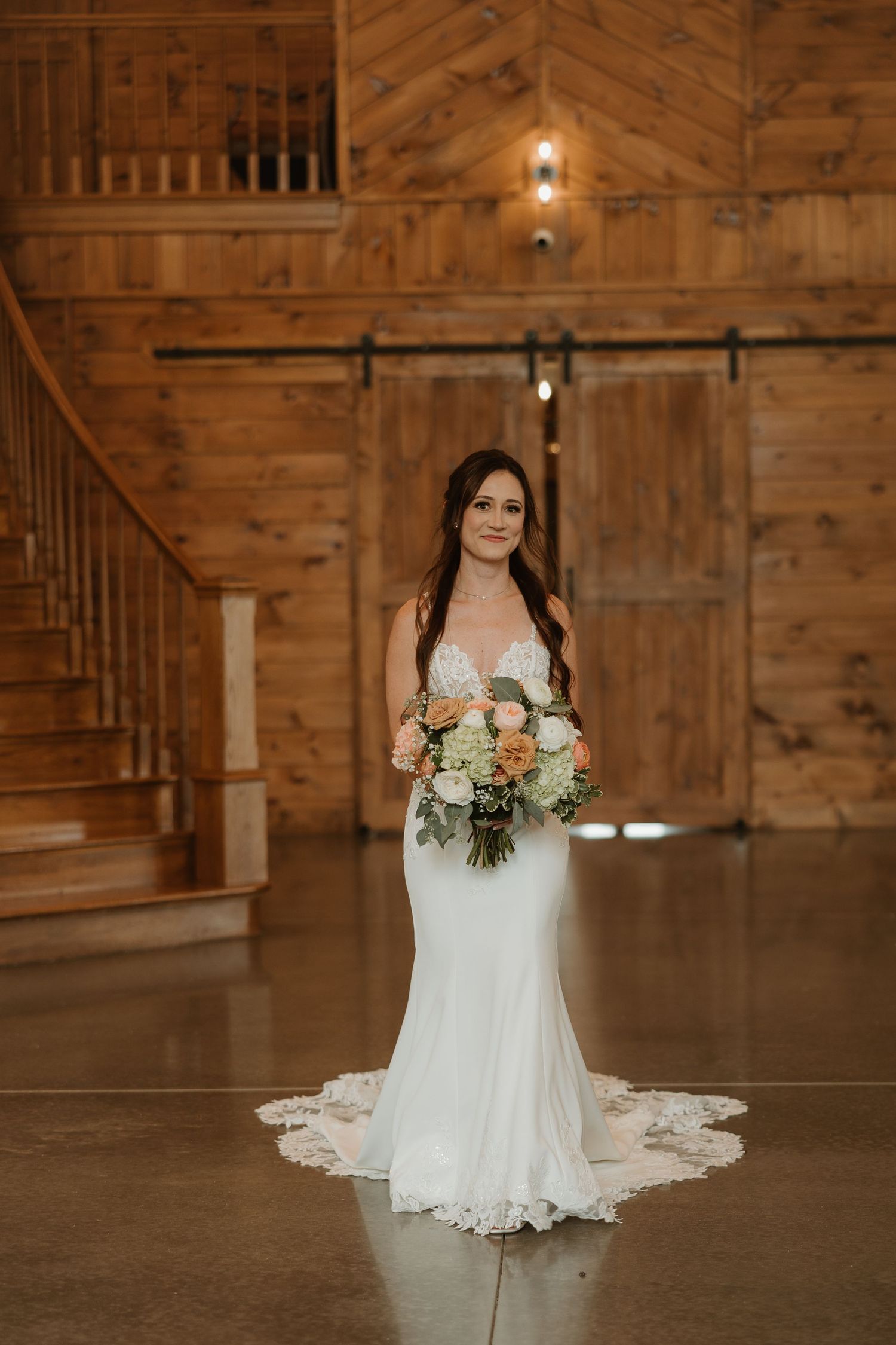 A rustic barn wedding venue interior with wooden walls, stairs, and a bride in a white lace gown holding a bouquet.