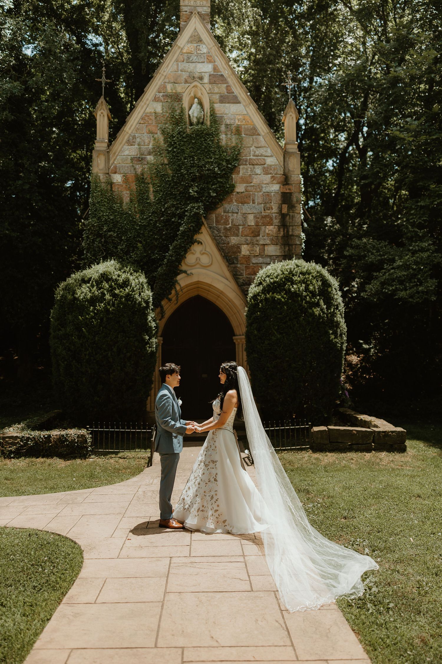 A couple stands together in front of a stone chapel with Gothic architecture and manicured shrubs on a sunny day.
