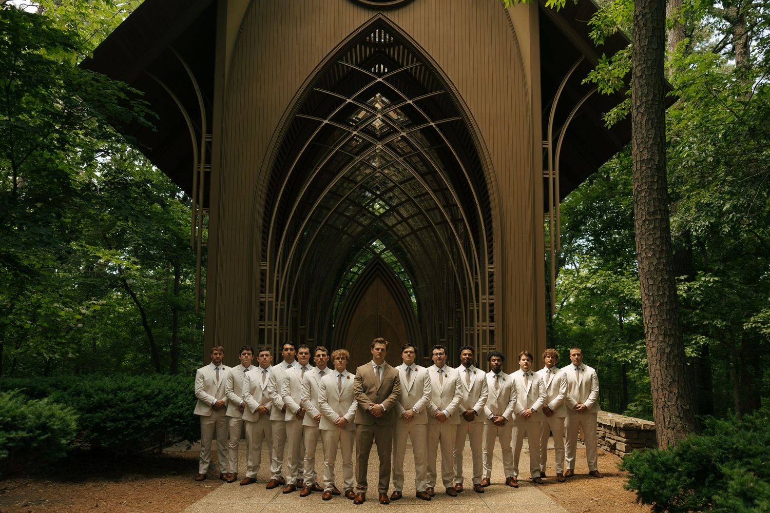 Groomsmen in tan suits standing under Gothic arched chapel canopy surrounded by lush green trees at outdoor wedding.