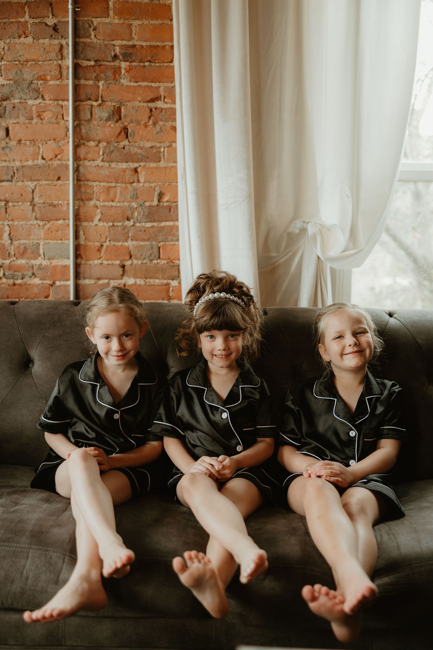 Three young girls in matching dark satin pajamas sitting barefoot on a gray tufted sofa near a brick wall.
