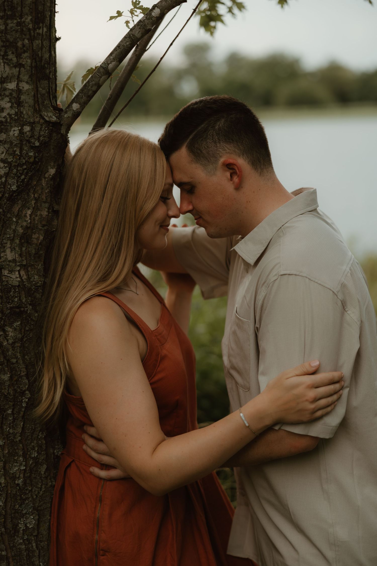 Couple sharing a romantic moment by a tree near a lake, woman in red dress and man in beige shirt embracing.