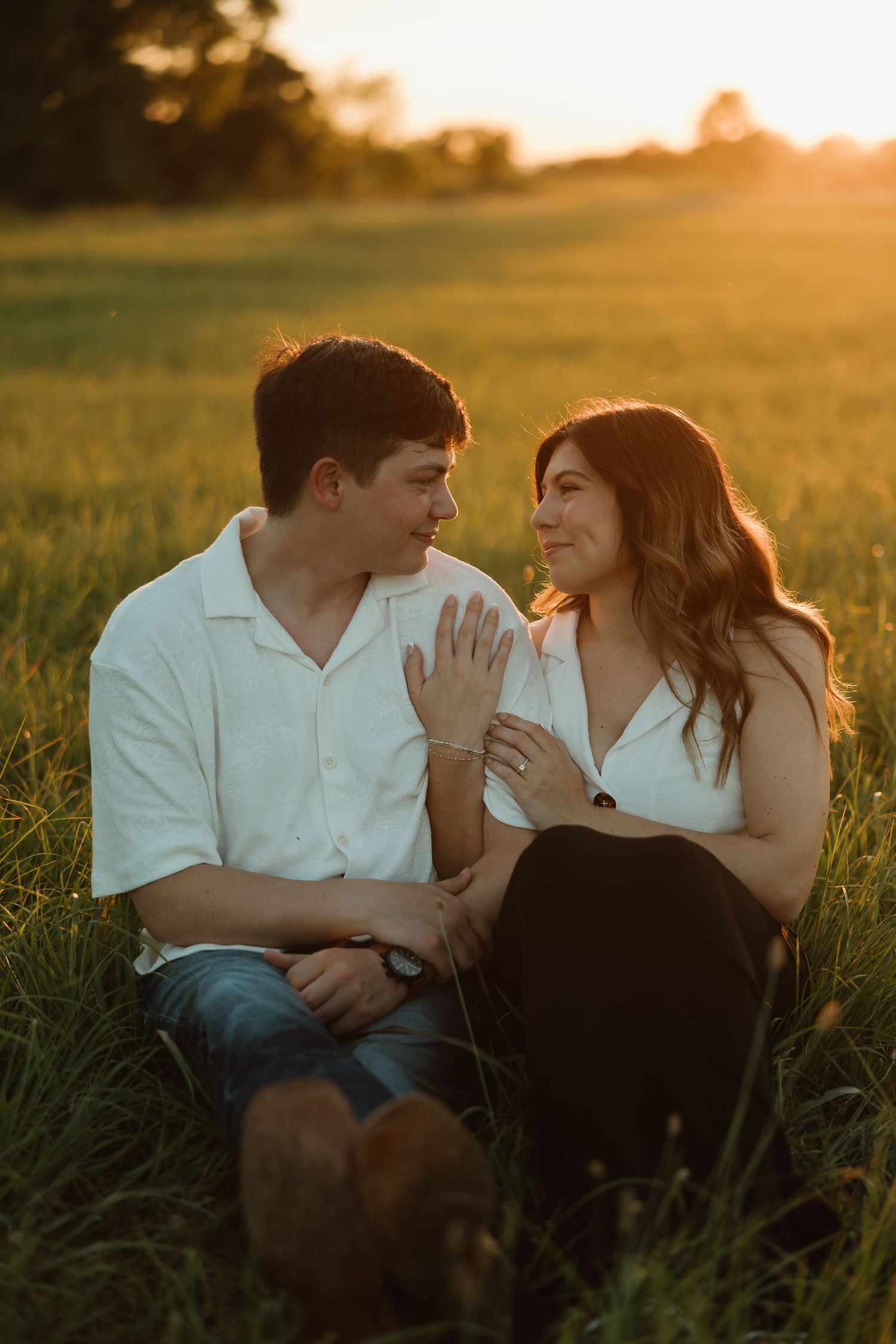 A romantic couple sits together in a grassy field at golden hour as sunlight creates a warm glow in the background.