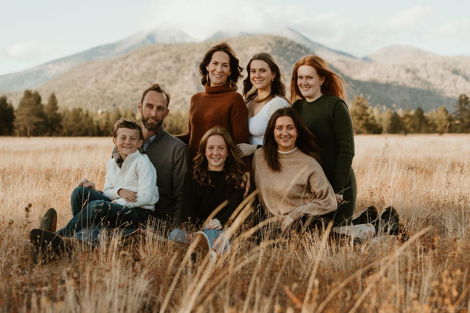 Group sitting together in a golden wheat field with mountain peaks and evergreen trees in the background at sunset.