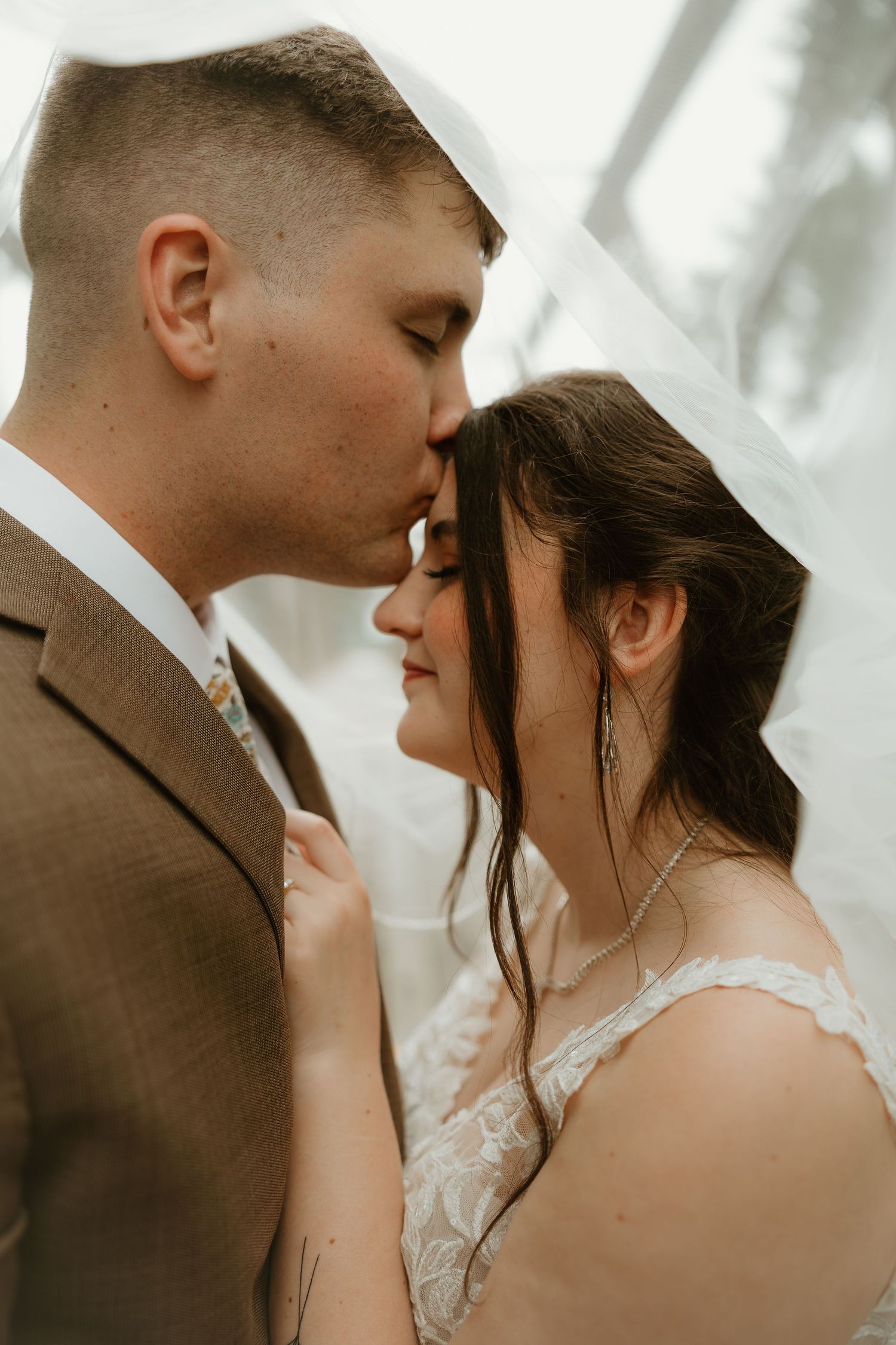 Intimate romantic wedding portrait in warm lighting shows tender forehead kiss between couple in brown suit and lace dress.