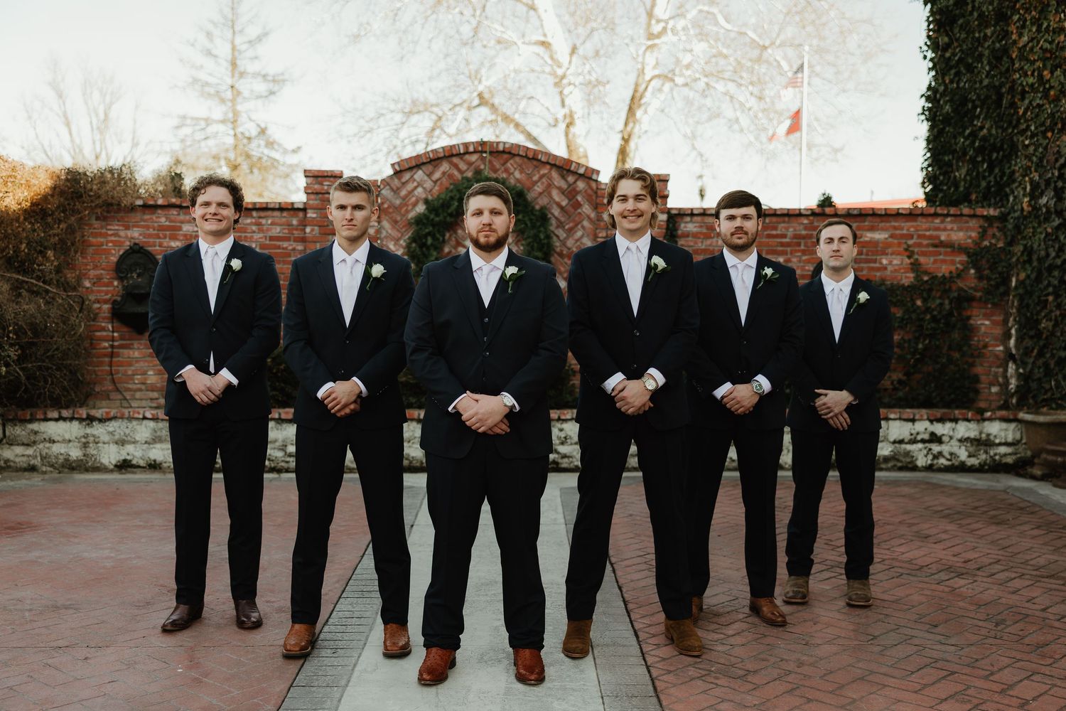 A wedding party in black suits and formal attire poses together outdoors against a brick wall.