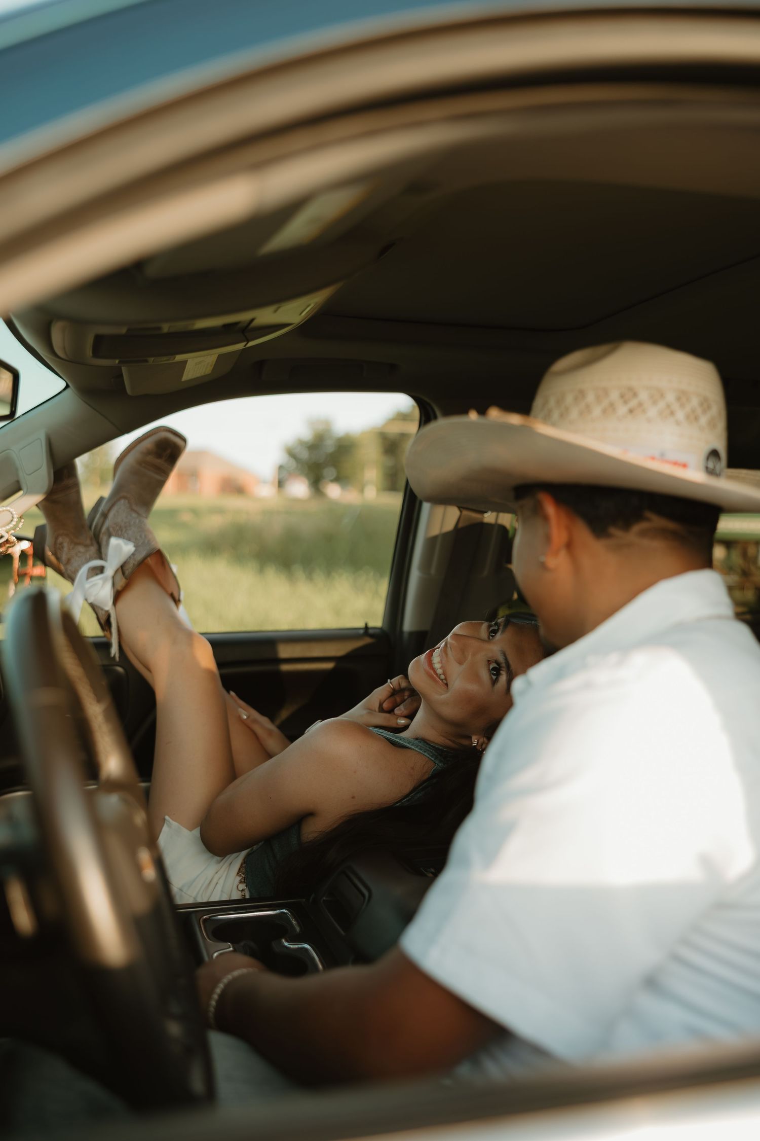 Couple in a truck cab, woman lying across seat smiling, man in cowboy hat, country lifestyle photo.