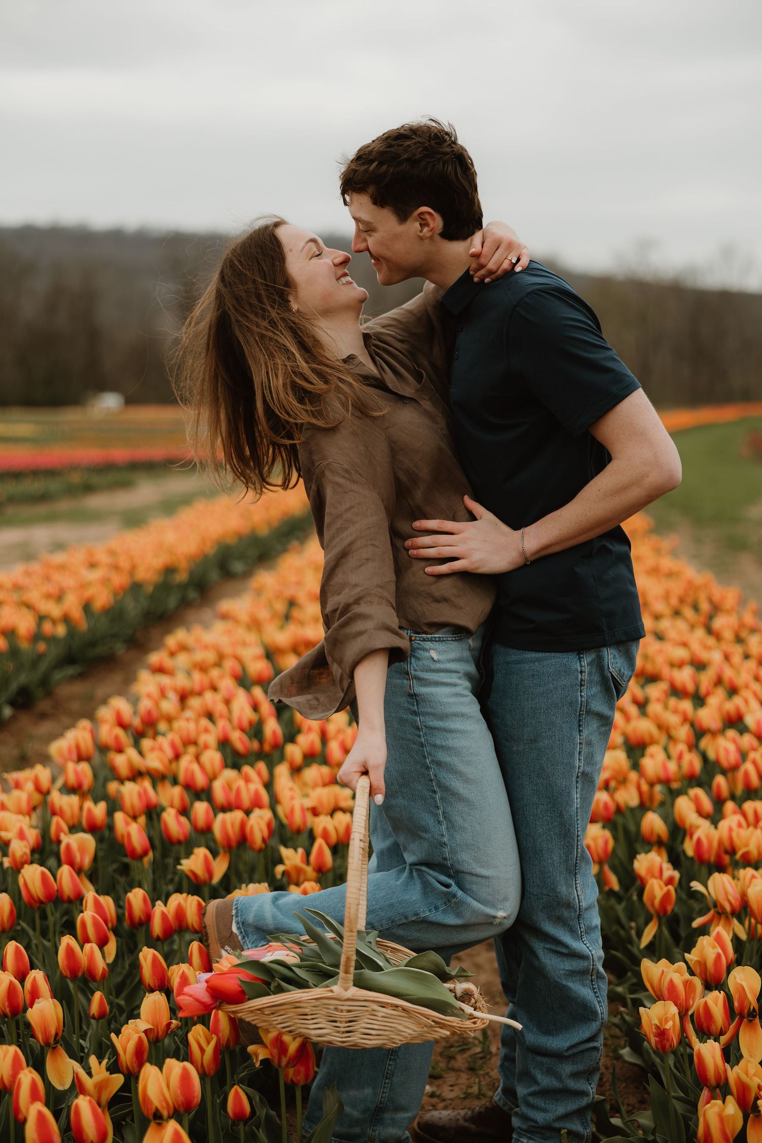 Couple embracing and laughing in a vibrant tulip field, woman holding a wicker basket of colorful flowers.