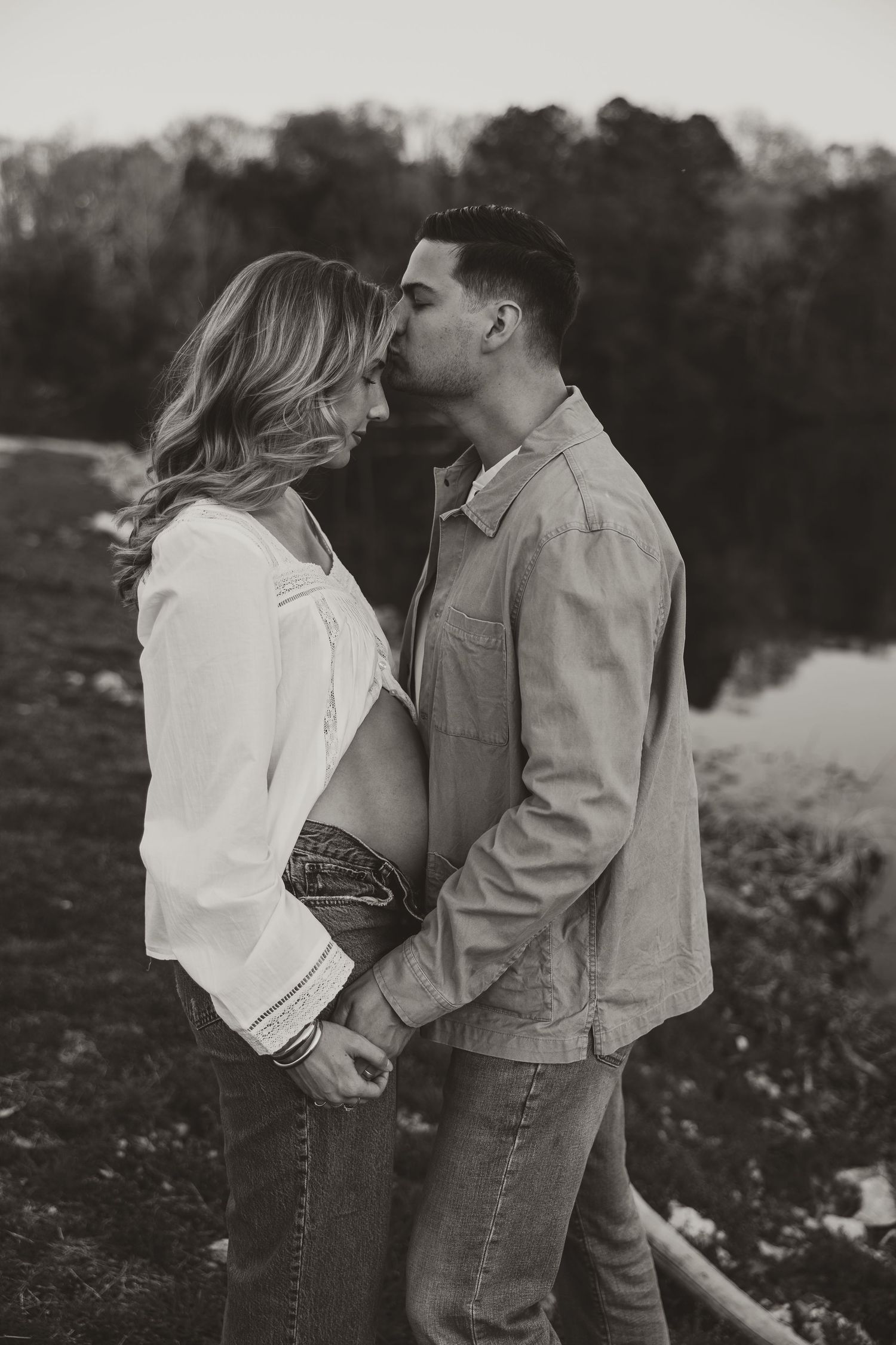 Black and white maternity photo of a couple holding hands near a pond, man kissing woman's forehead outdoors.