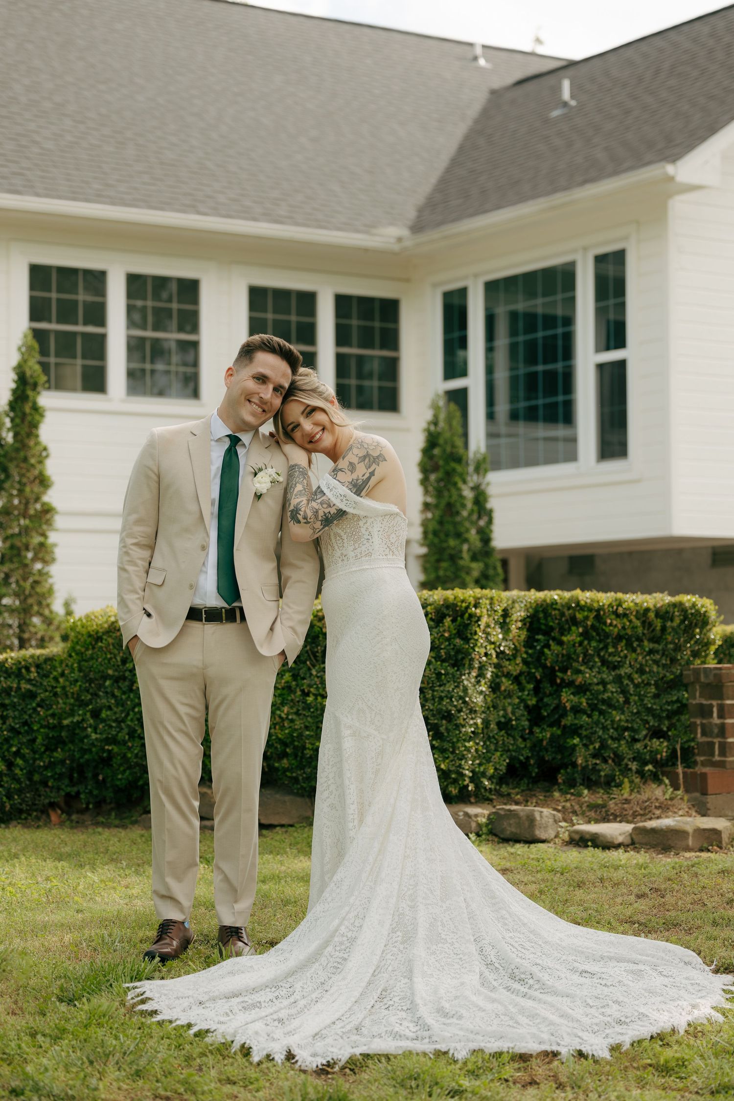 A bride in a lace gown and groom in a tan suit pose together on a lawn outside a white house.
