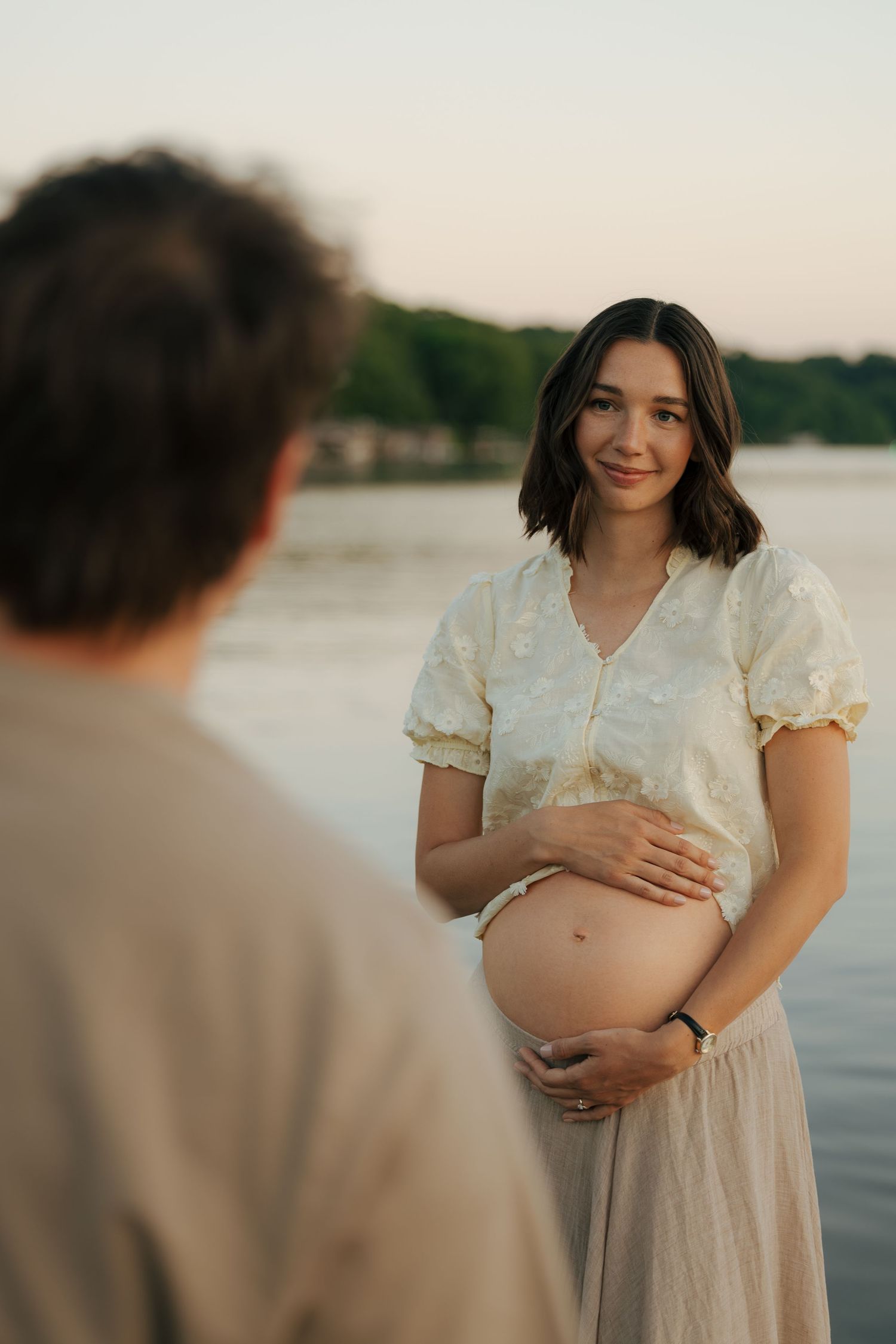 Pregnant woman in floral top cradling baby bump near a lake at sunset, smiling warmly at her partner.