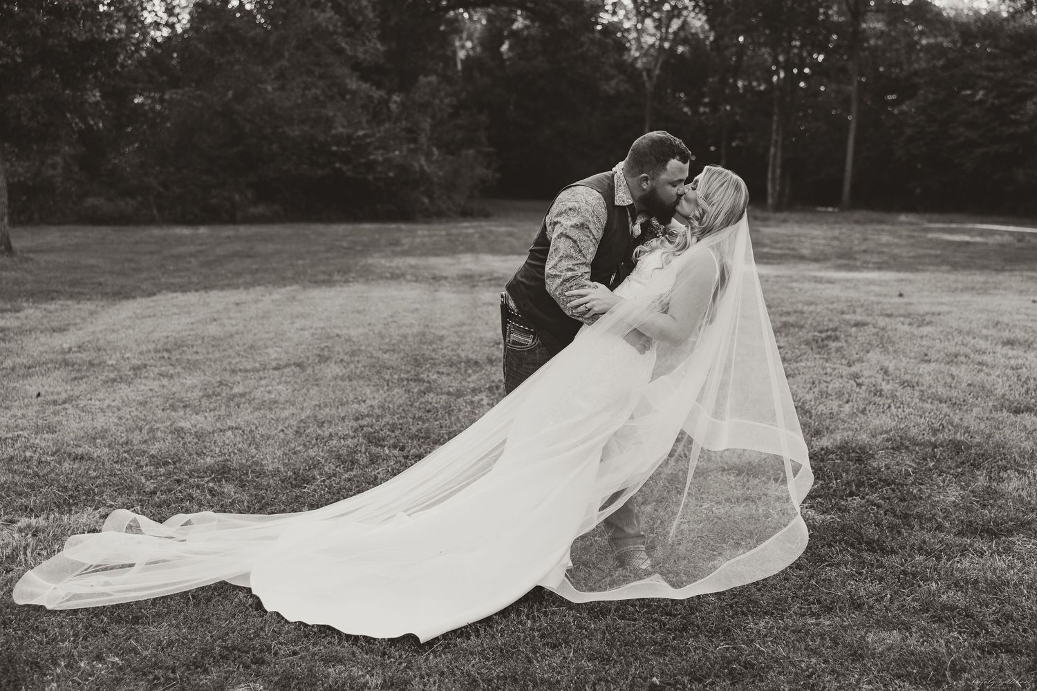 A romantic black and white wedding photo shows a couple embracing in a grassy field with a flowing veil in the wind.