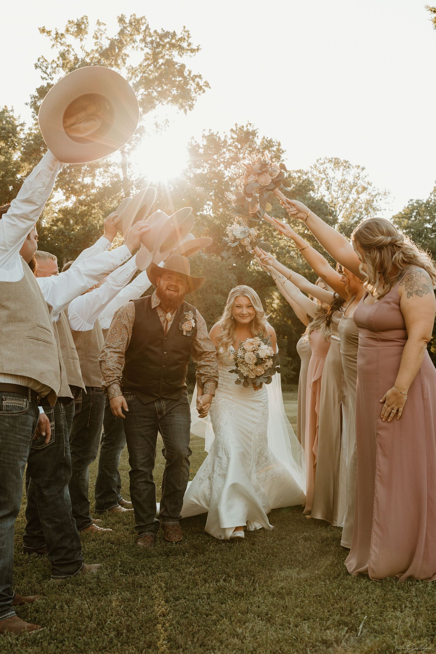 Wedding party celebrates outdoors at sunset with cowboy hat raised as newlyweds walk through on grass.