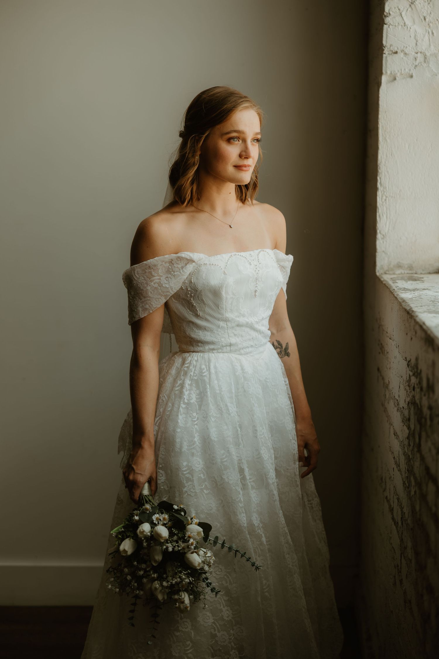 A bride in an off-shoulder white lace wedding gown poses with a dark floral bouquet against a textured wall.