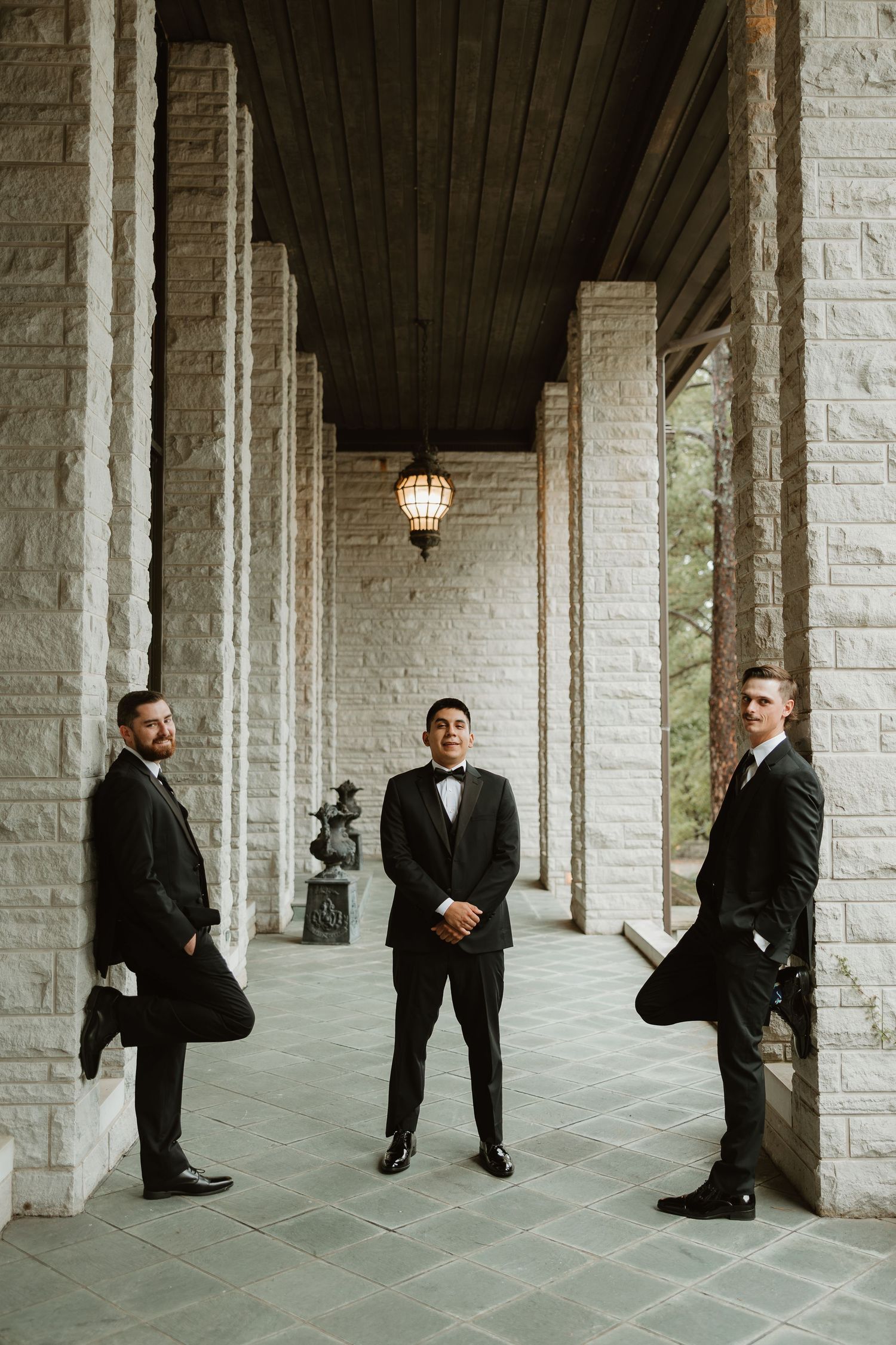 Groomsmen in black suits pose creatively between white stone columns in an elegant covered walkway.