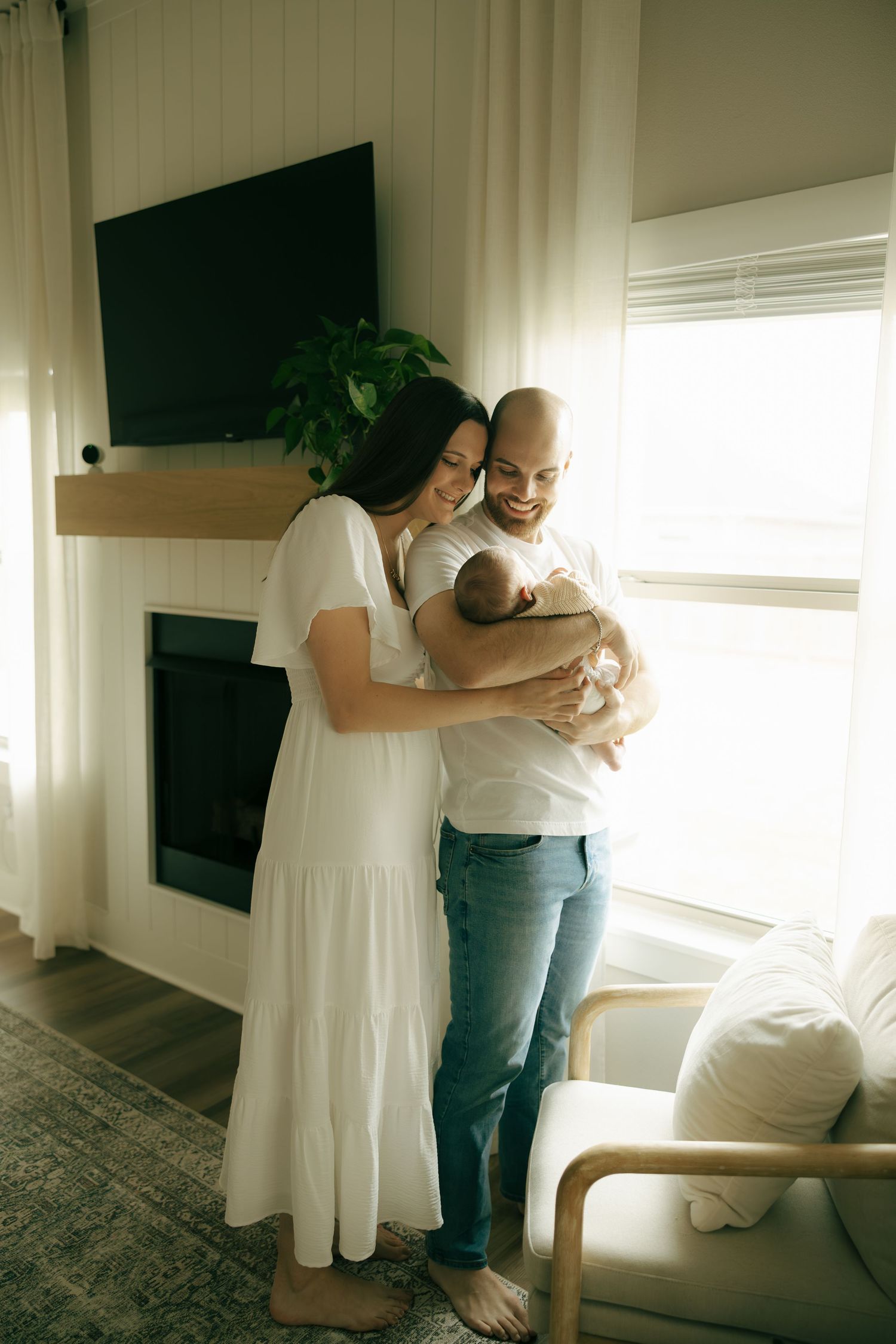 Happy couple in white outfits cradling newborn baby by fireplace in bright modern living room.