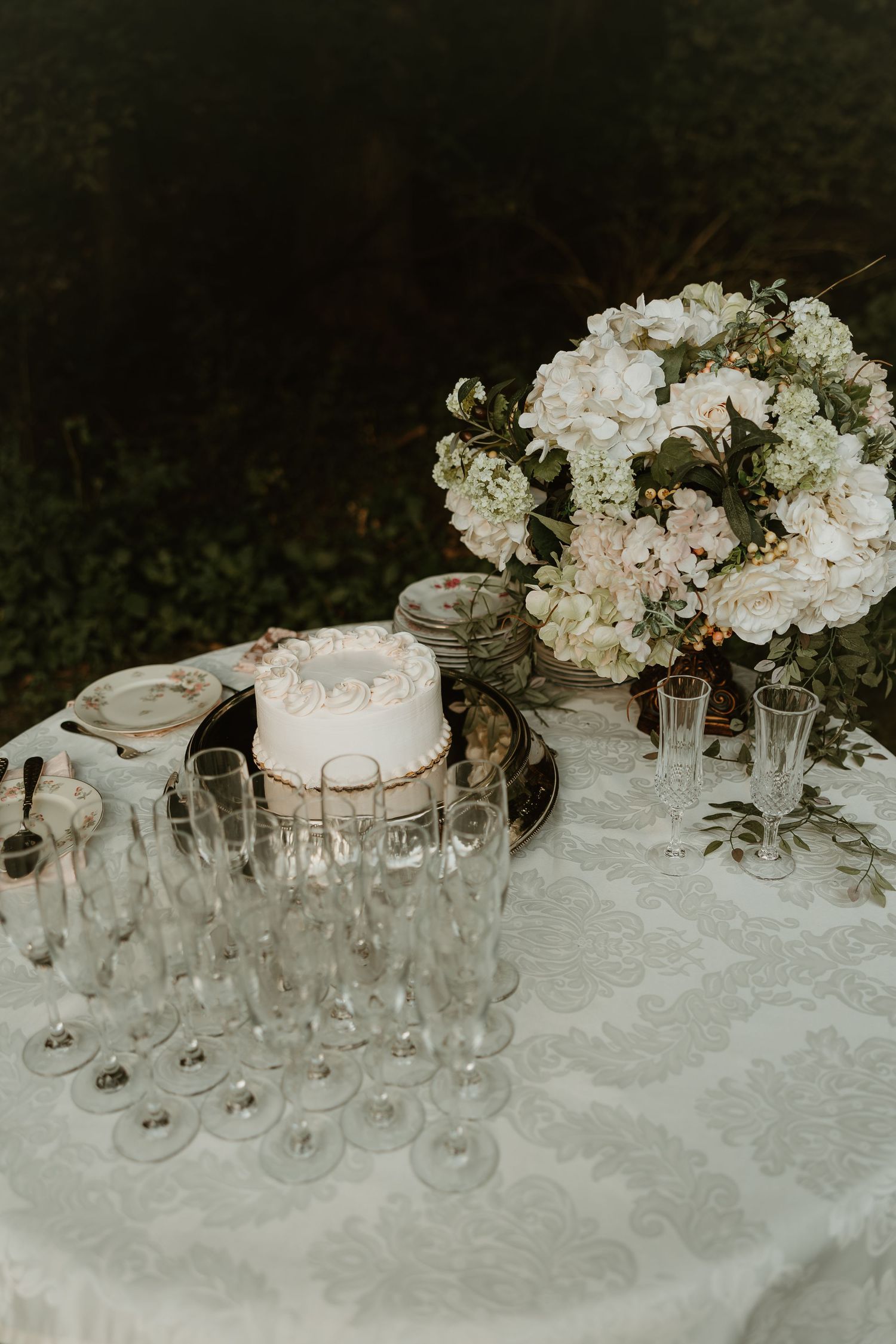 Elegant wedding table setting with white floral centerpiece, champagne glasses and white cake on lace tablecloth.