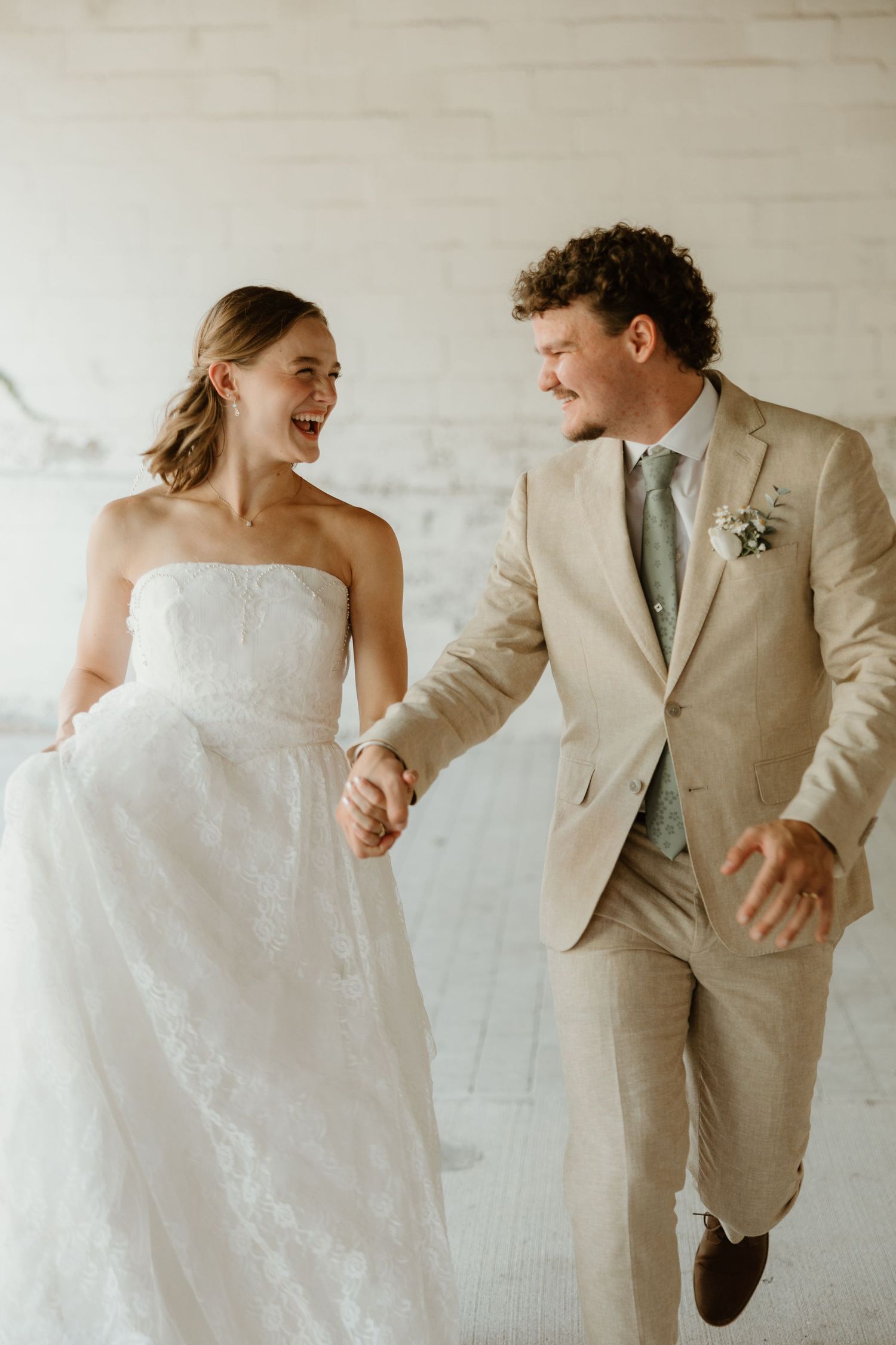 A joyful couple in wedding attire runs and laughs together against a white wall during their wedding celebration.