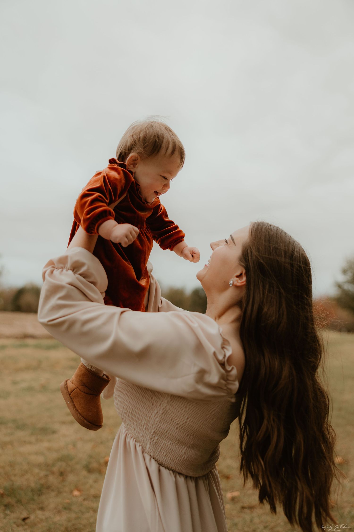 Mother in cream dress lifts baby wearing orange sweater in autumn field during golden hour moment.