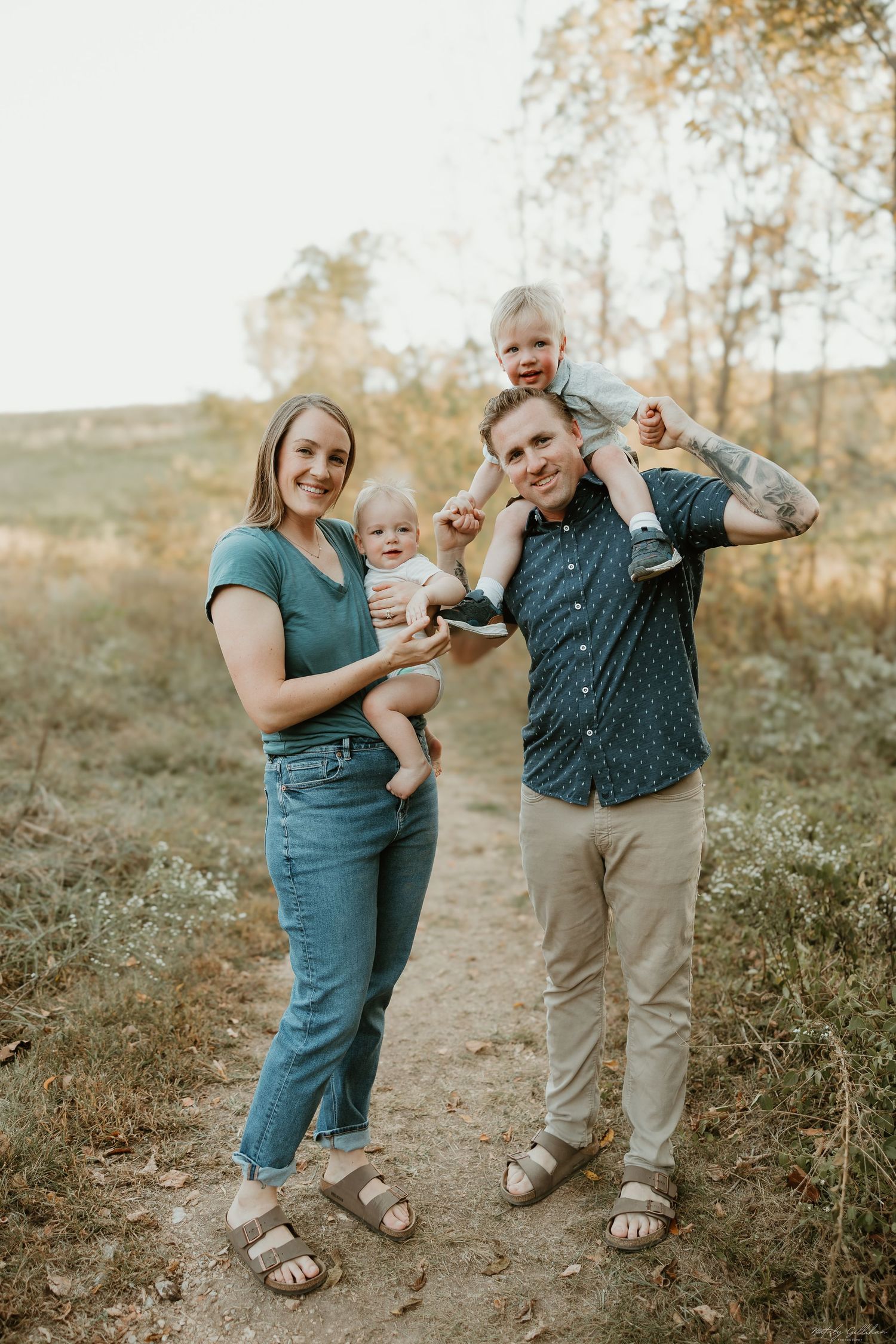 A family stands together on a dirt path during golden hour in a natural outdoor setting wearing casual attire.