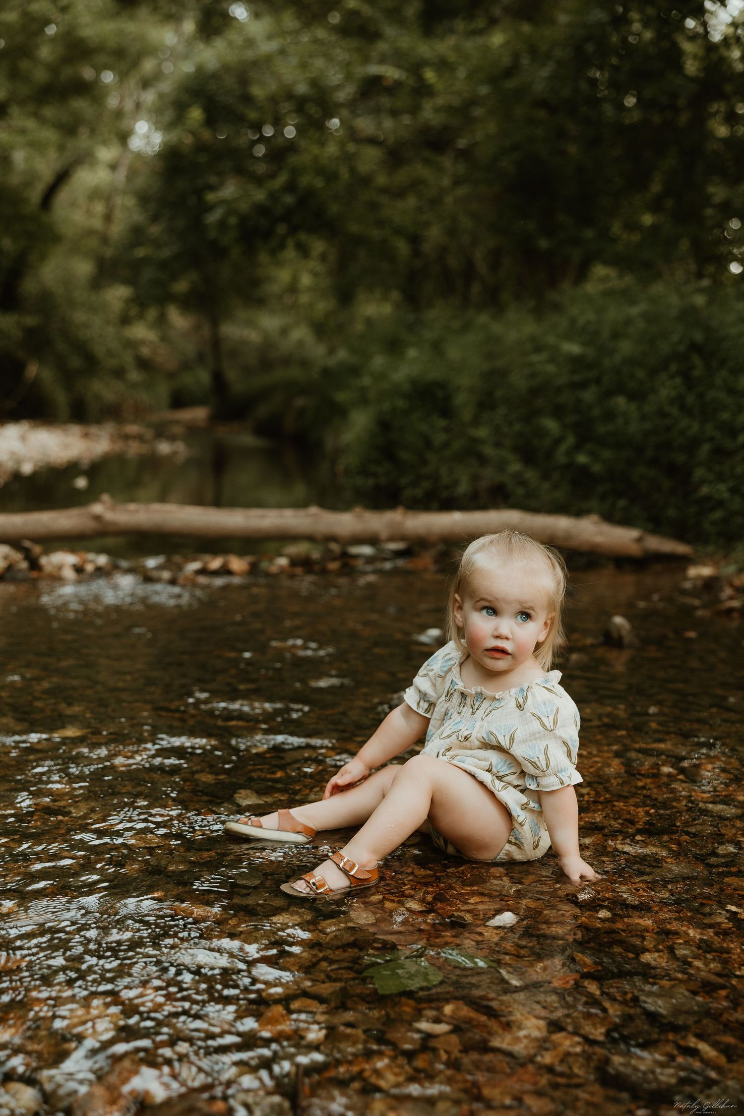 Small child sitting in a shallow creek with rocks and forest scenery in the background during summer.