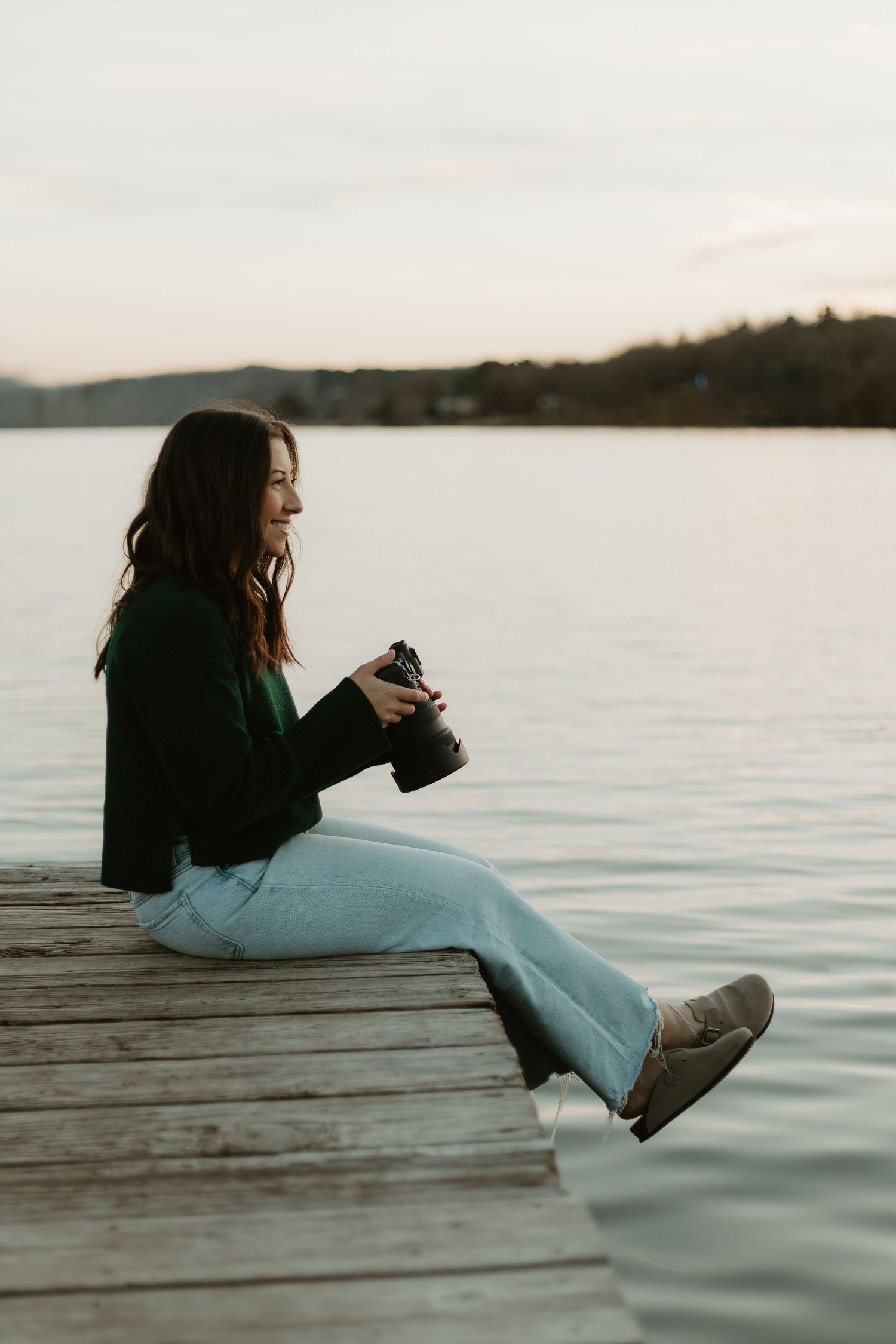 Woman in green sweater and jeans sits on wooden dock holding camera, smiling at serene lake during sunset.