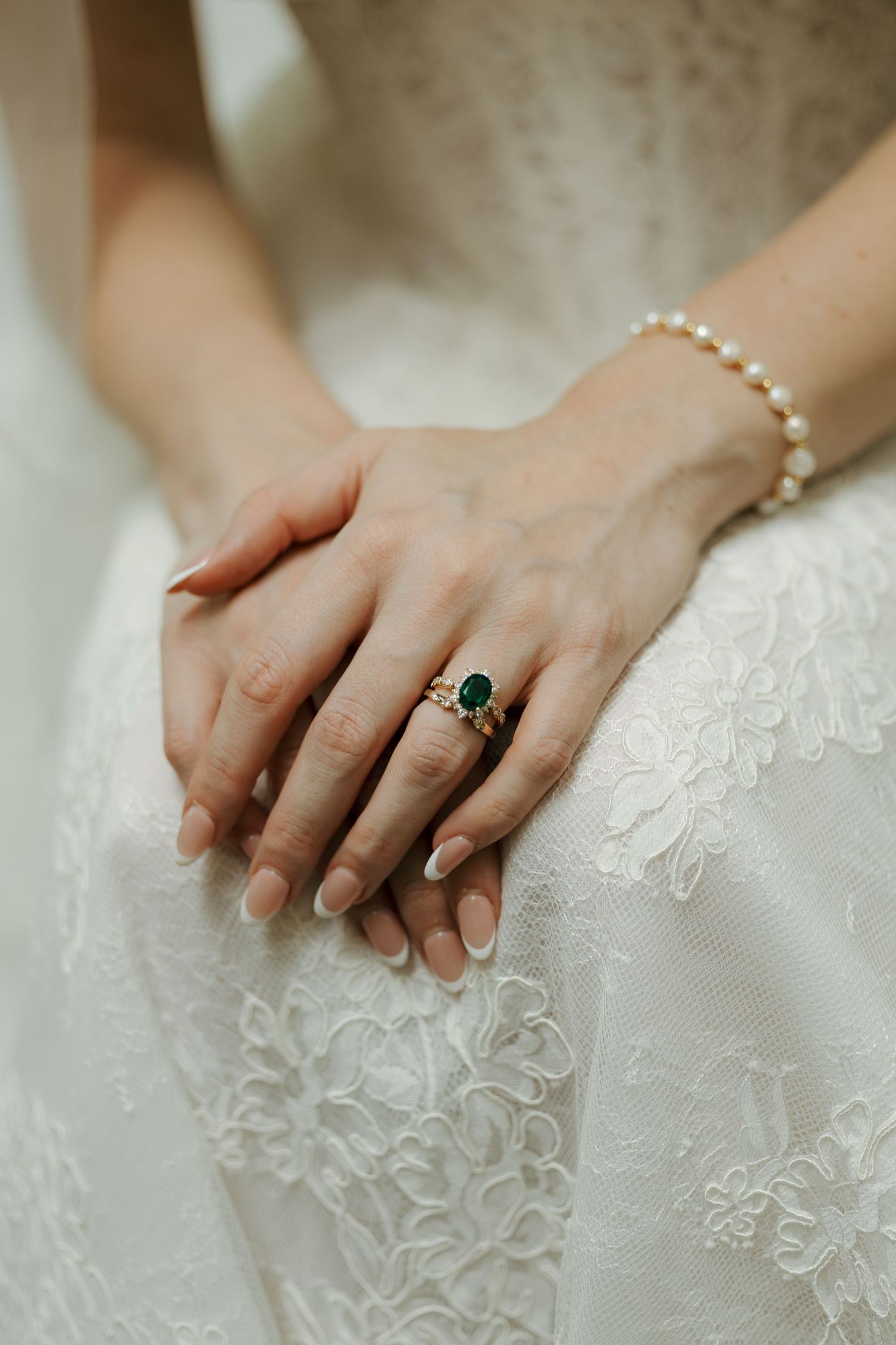 Bride's hands resting on lace wedding dress, wearing emerald green gemstone ring and pearl bracelet.