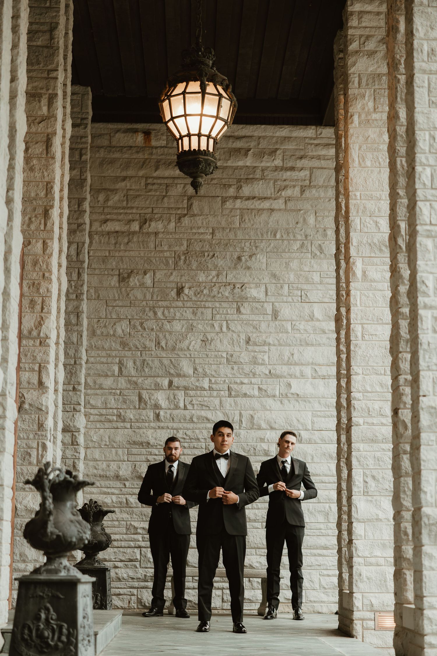 Groomsmen in black suits pose playfully against stone pillars and walls at an elegant venue with hanging lanterns.