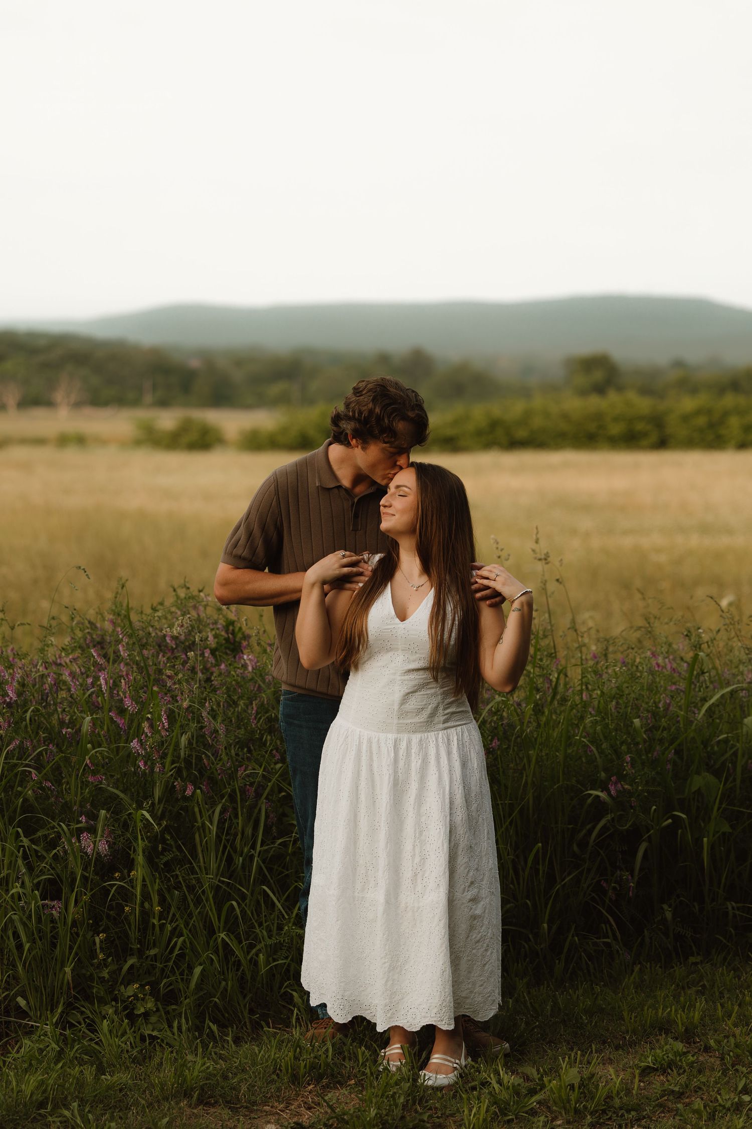 A couple shares a tender moment in a golden meadow, man kissing womans head as she wears a white dress.