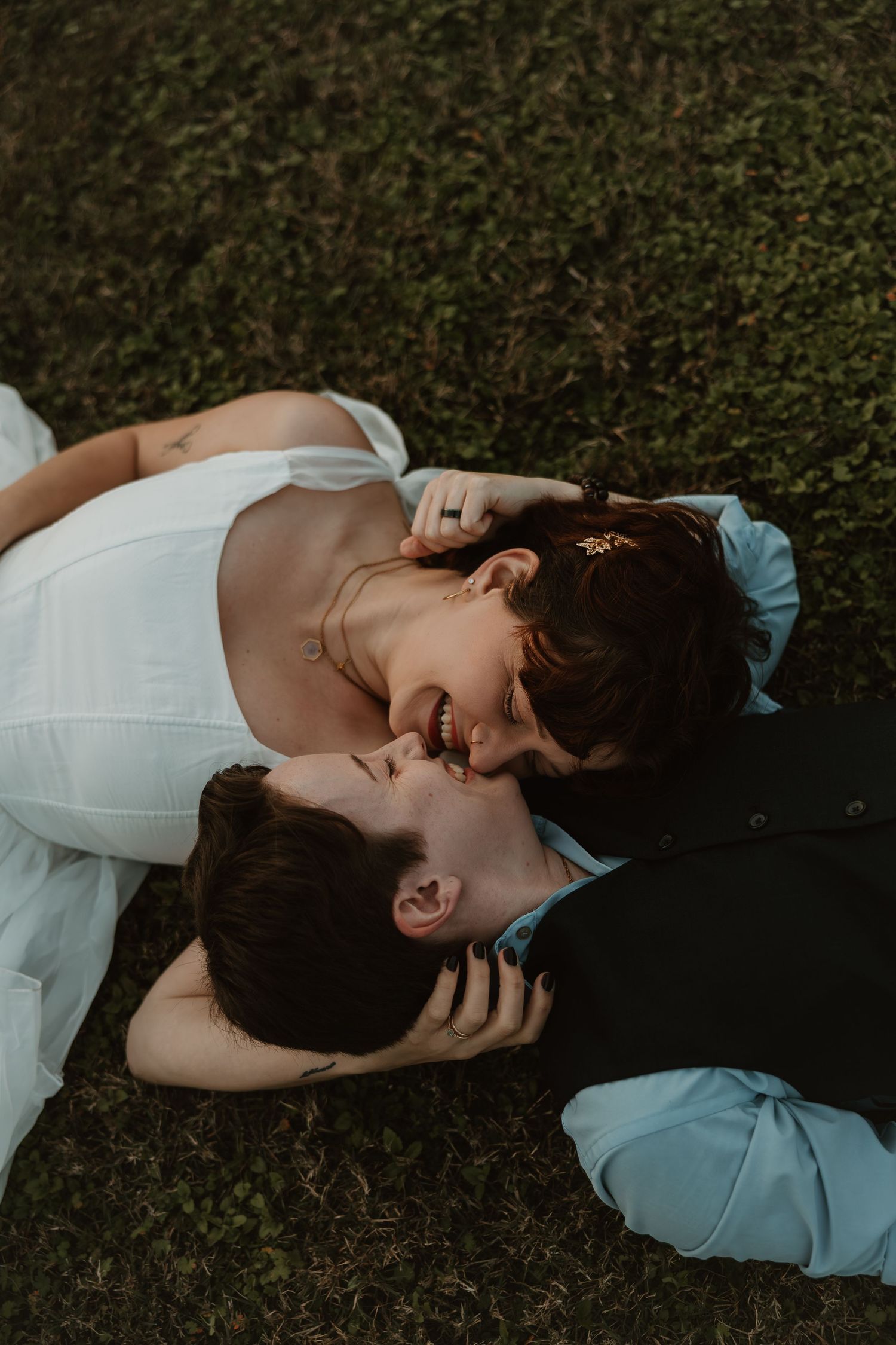 A bride and groom lying on green grass, sharing a romantic kiss in their wedding attire.
