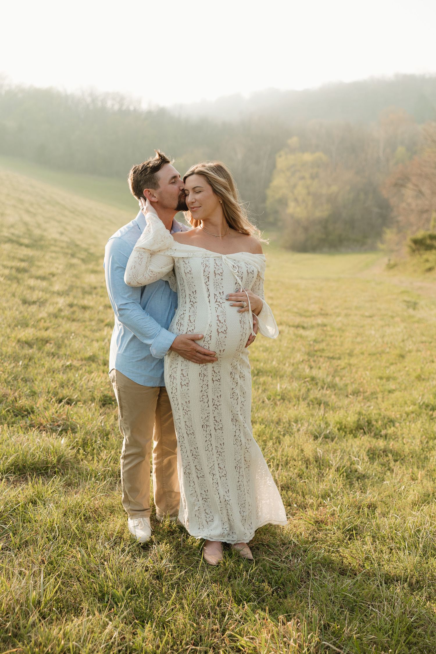 A couple shares a tender kiss in a sunlit field, the woman in a white lace maternity gown cradling her baby bump.