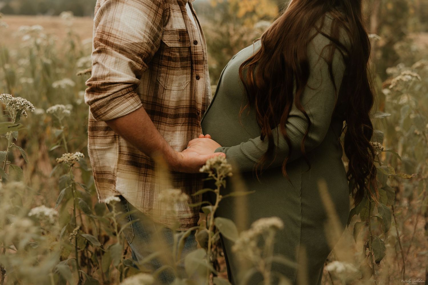 Two people hold hands while standing in a field of tall wildflowers and grass at sunset.