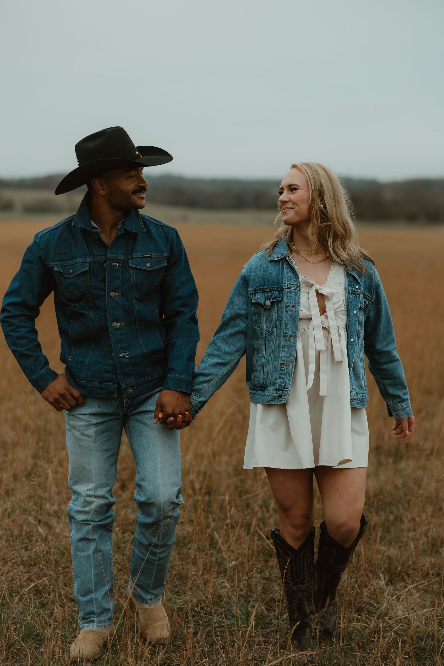 A couple in western attire stands together in a golden field at sunset, wearing denim jackets and cowboy boots.