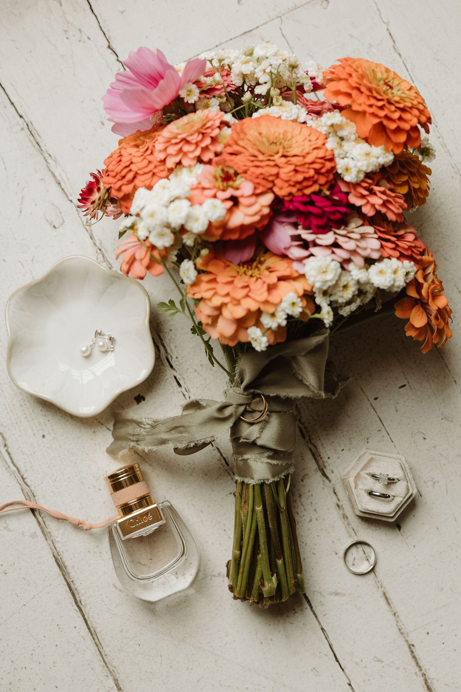 Bridal flat lay with orange and pink zinnia bouquet, perfume bottle, wedding rings, and earrings on white wood.