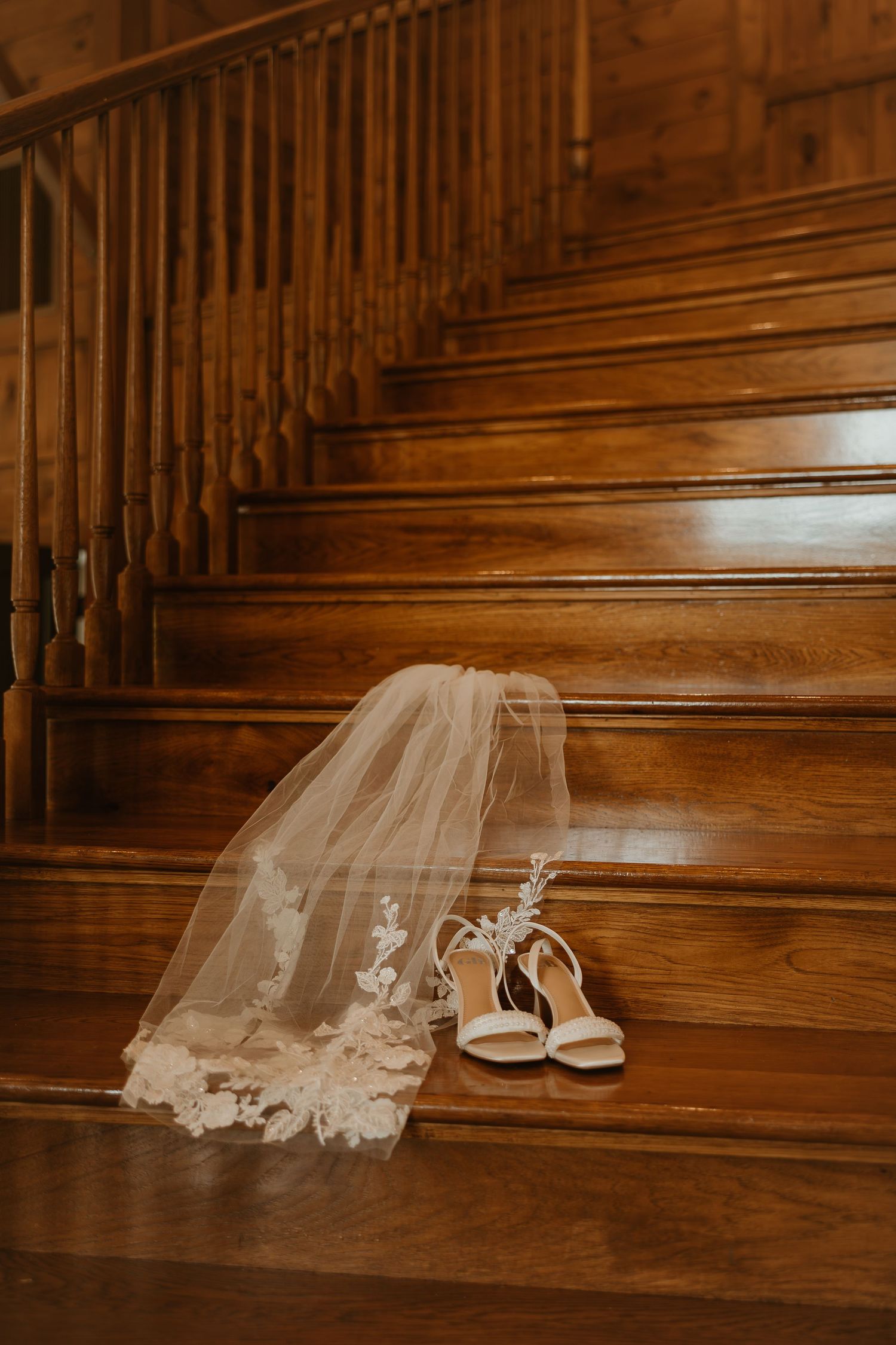 A wedding veil and white bridal shoes rest on polished wooden stairs in a warm sepia-toned photograph.