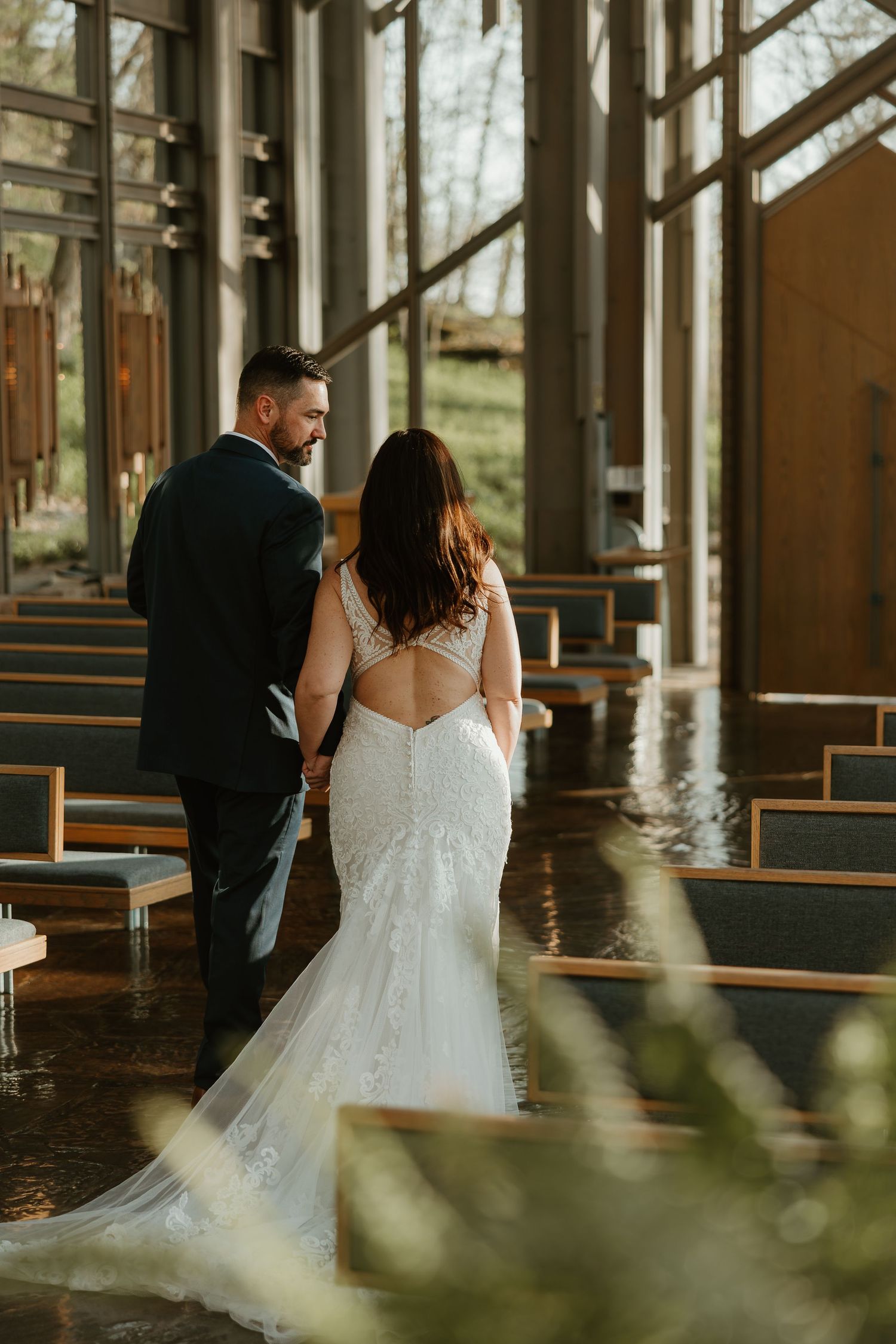 A couple in wedding attire standing together inside a modern chapel with large windows and wooden pews.