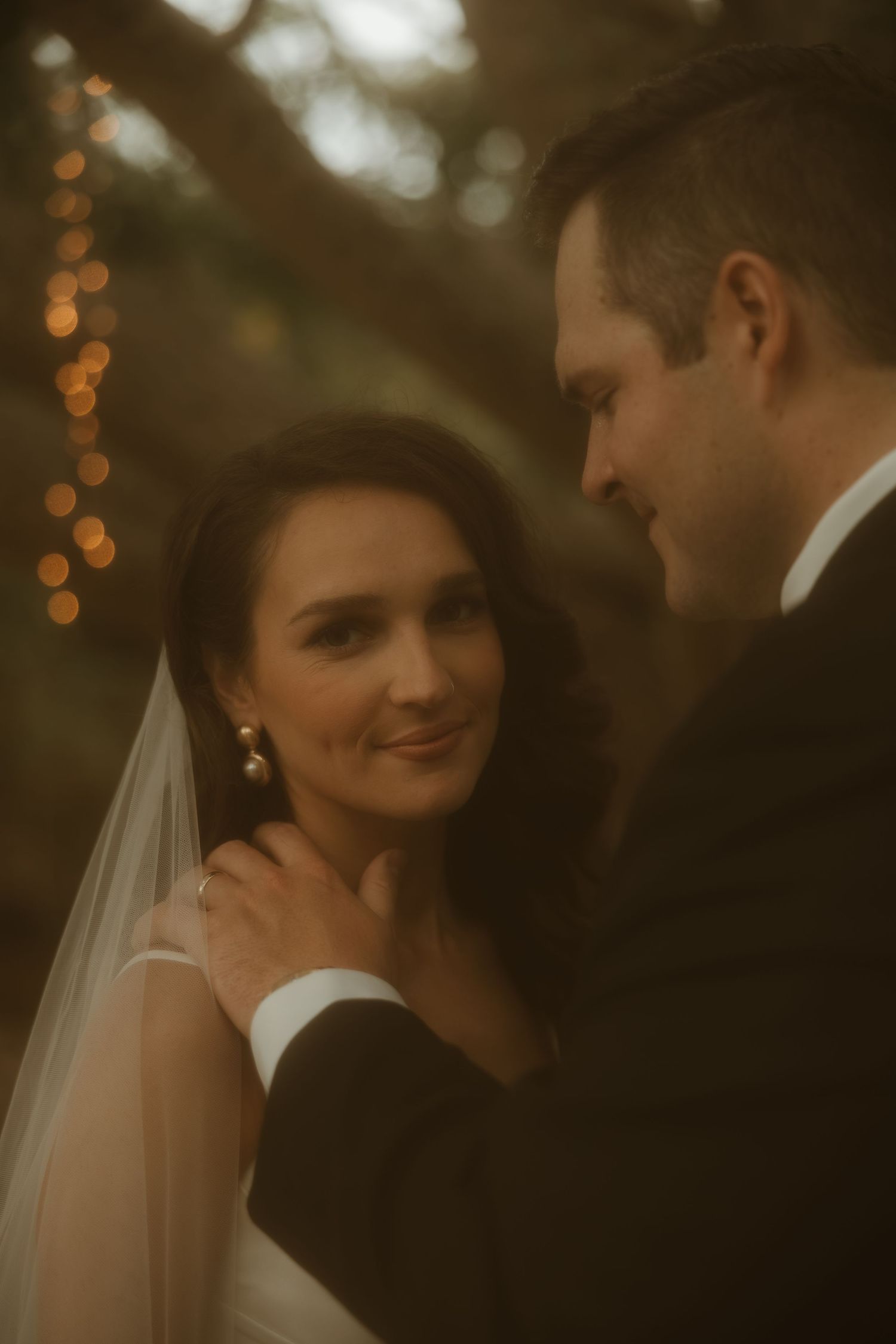 A bride in a white veil smiles warmly at her groom in a suit, surrounded by soft bokeh lights in an outdoor setting.