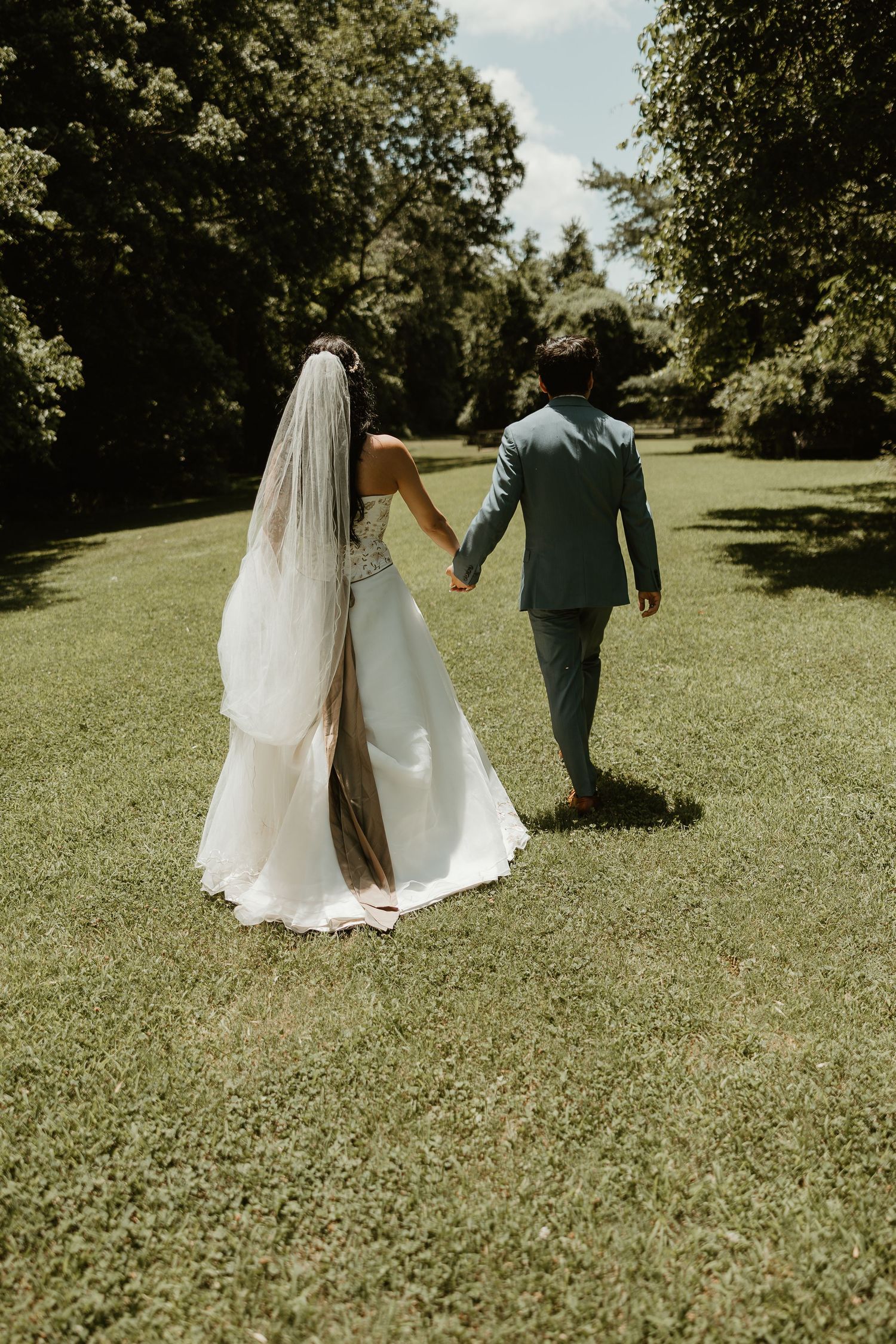 A couple in wedding attire holding hands while walking together through a lush green garden setting.