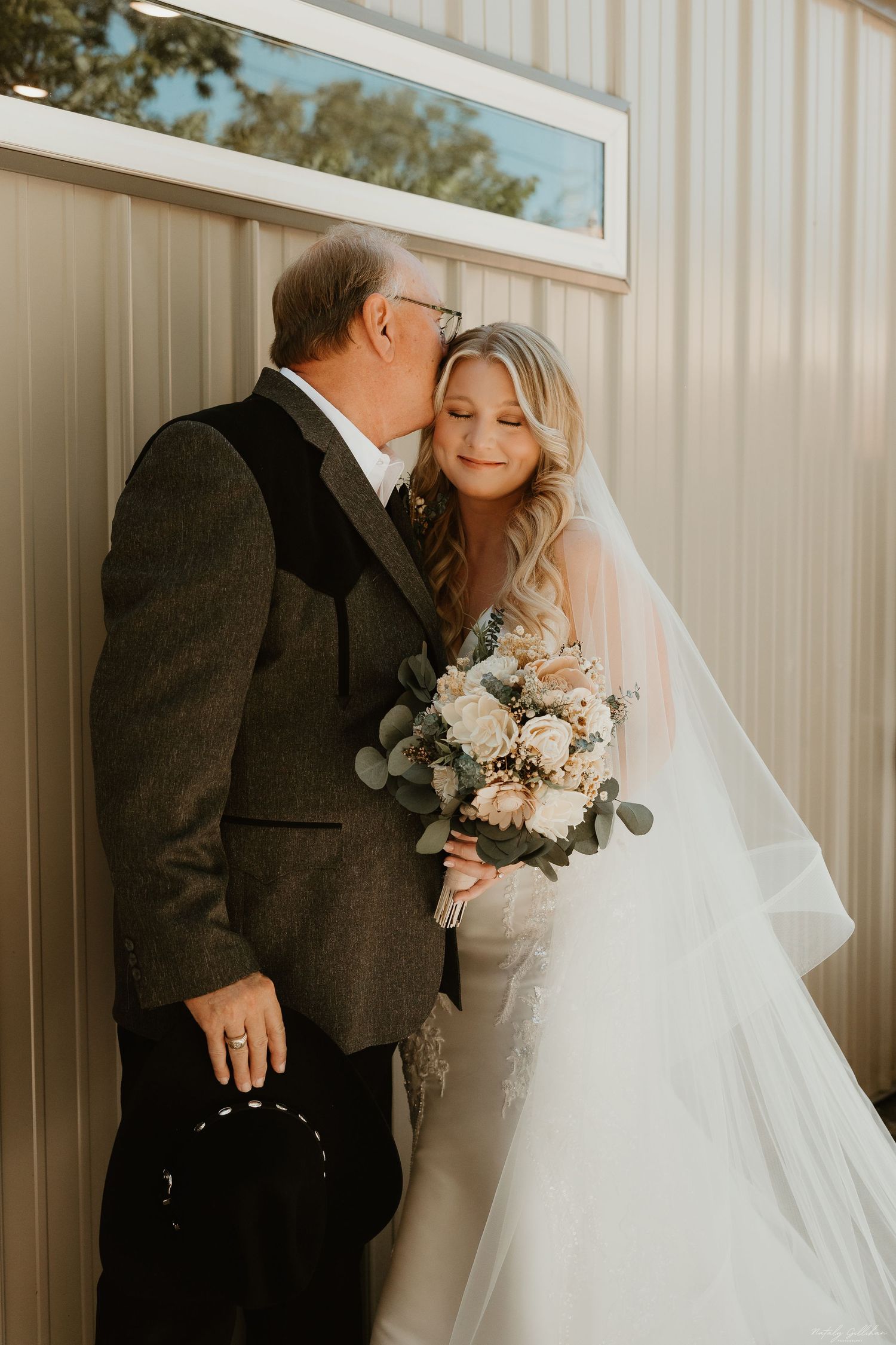A tender wedding day moment between a bride in white dress holding a neutral bouquet and someone in a dark suit.