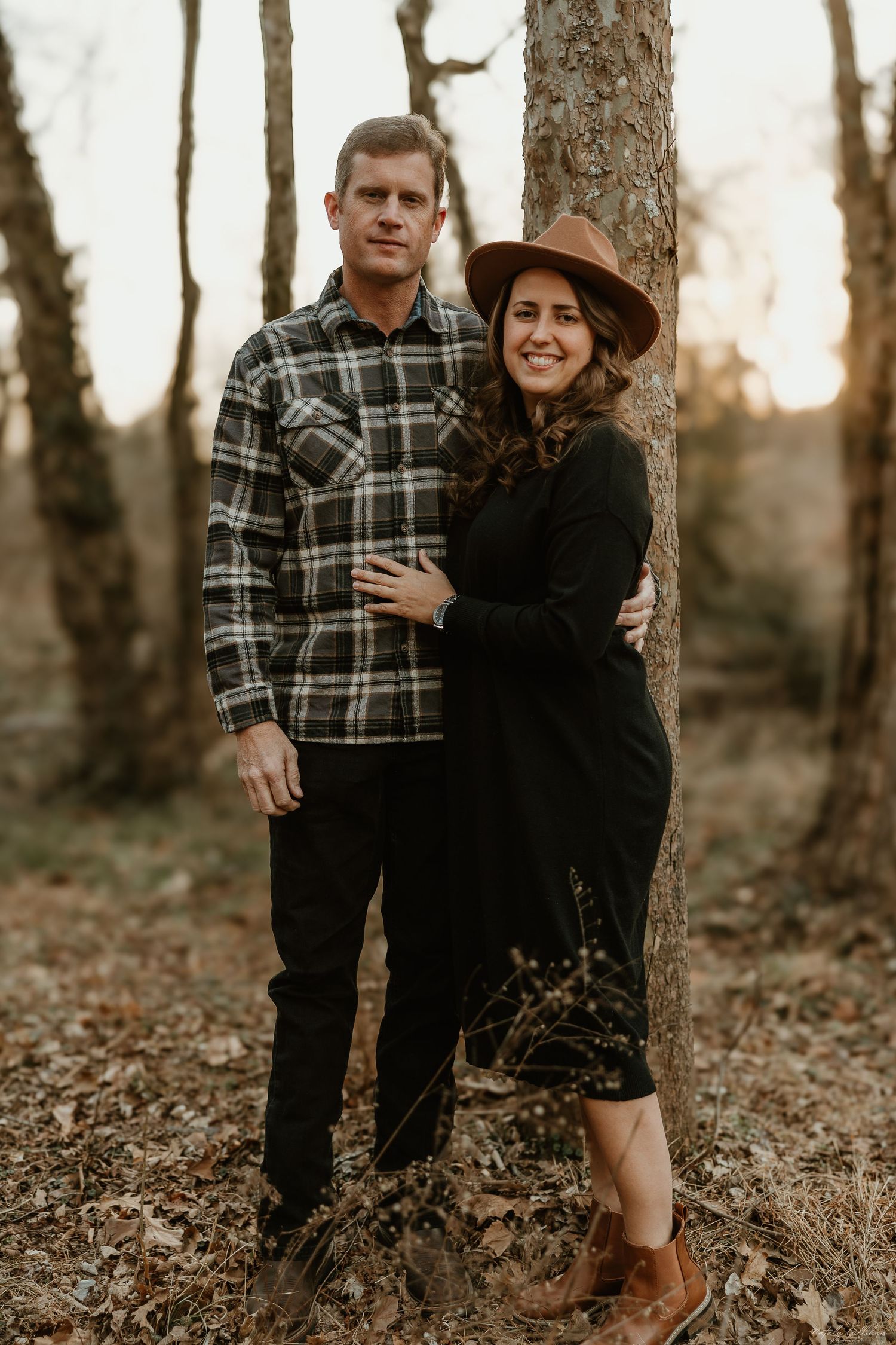 Romantic forest portrait featuring plaid shirt and black dress with brown fedora hat.