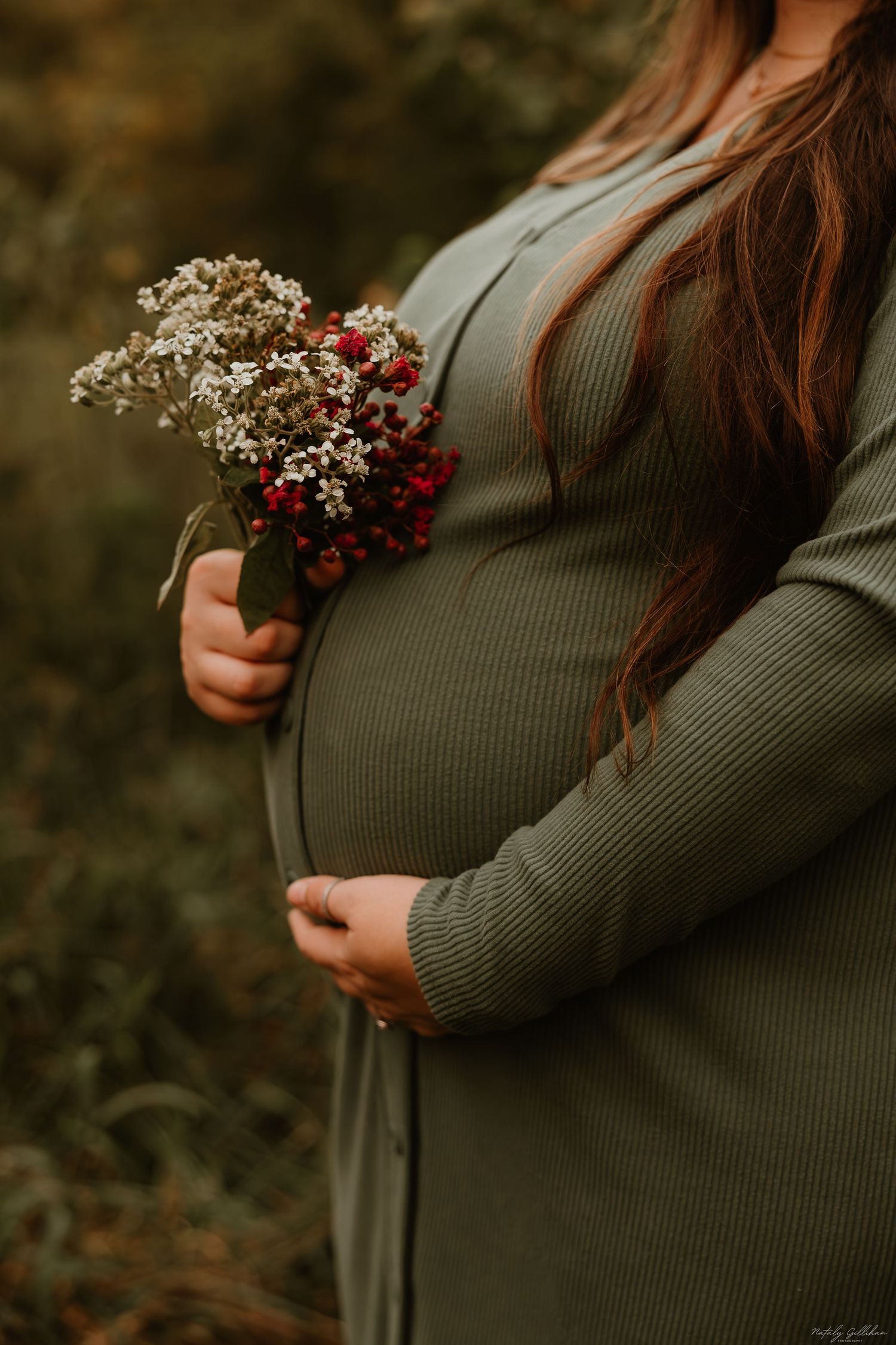 A pregnant belly in a green sweater dress is cradled while holding delicate white and red flowers in autumn.