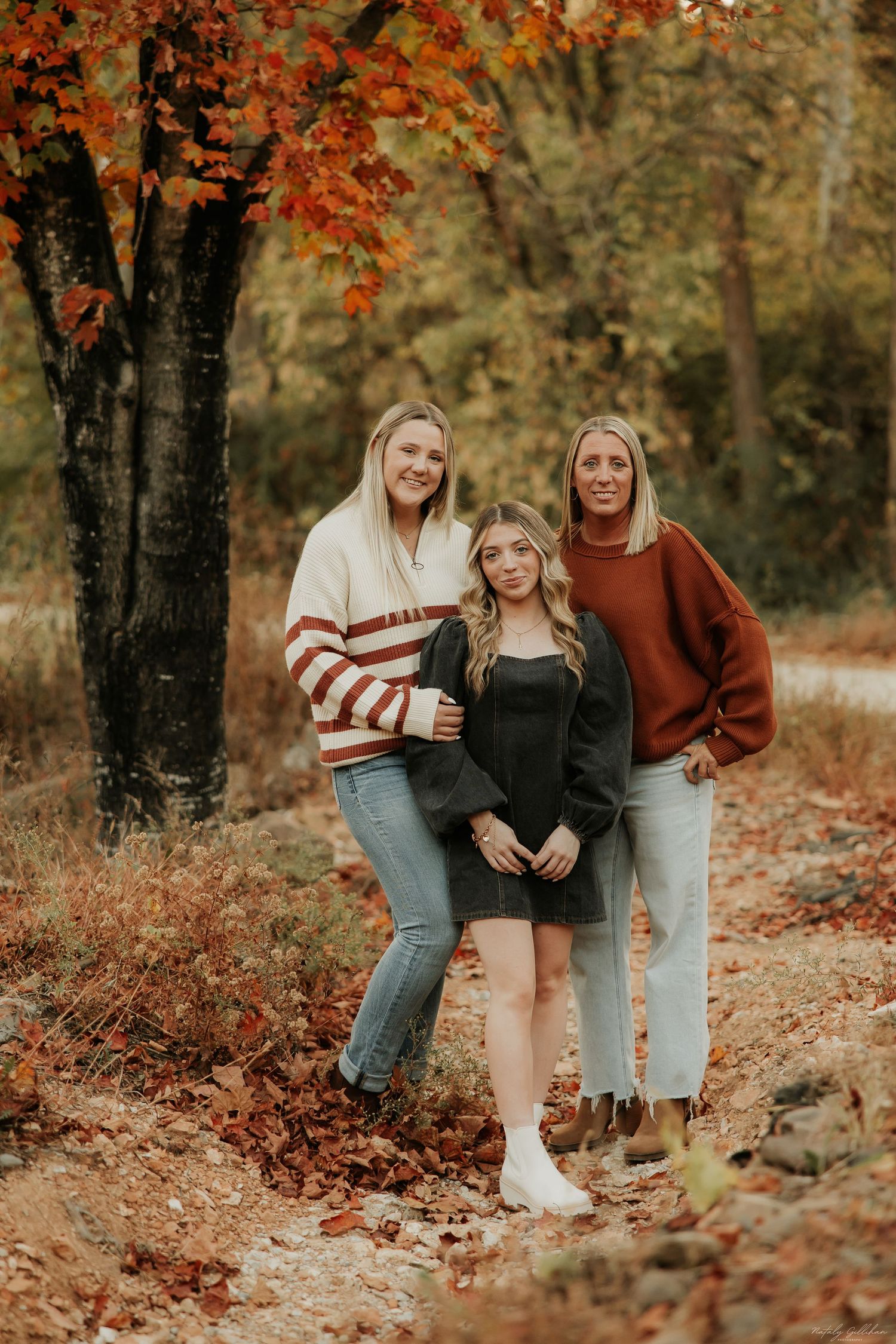 Three friends pose together on a woodland trail surrounded by fall foliage and orange autumn leaves.