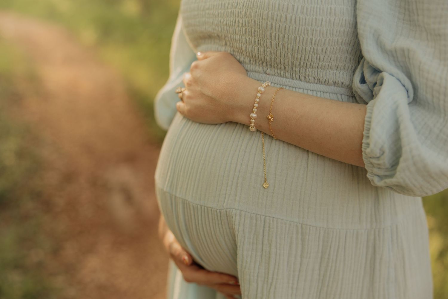 Close-up of pregnant belly in soft beige dress with delicate bracelet-adorned hand resting on bump at sunset.